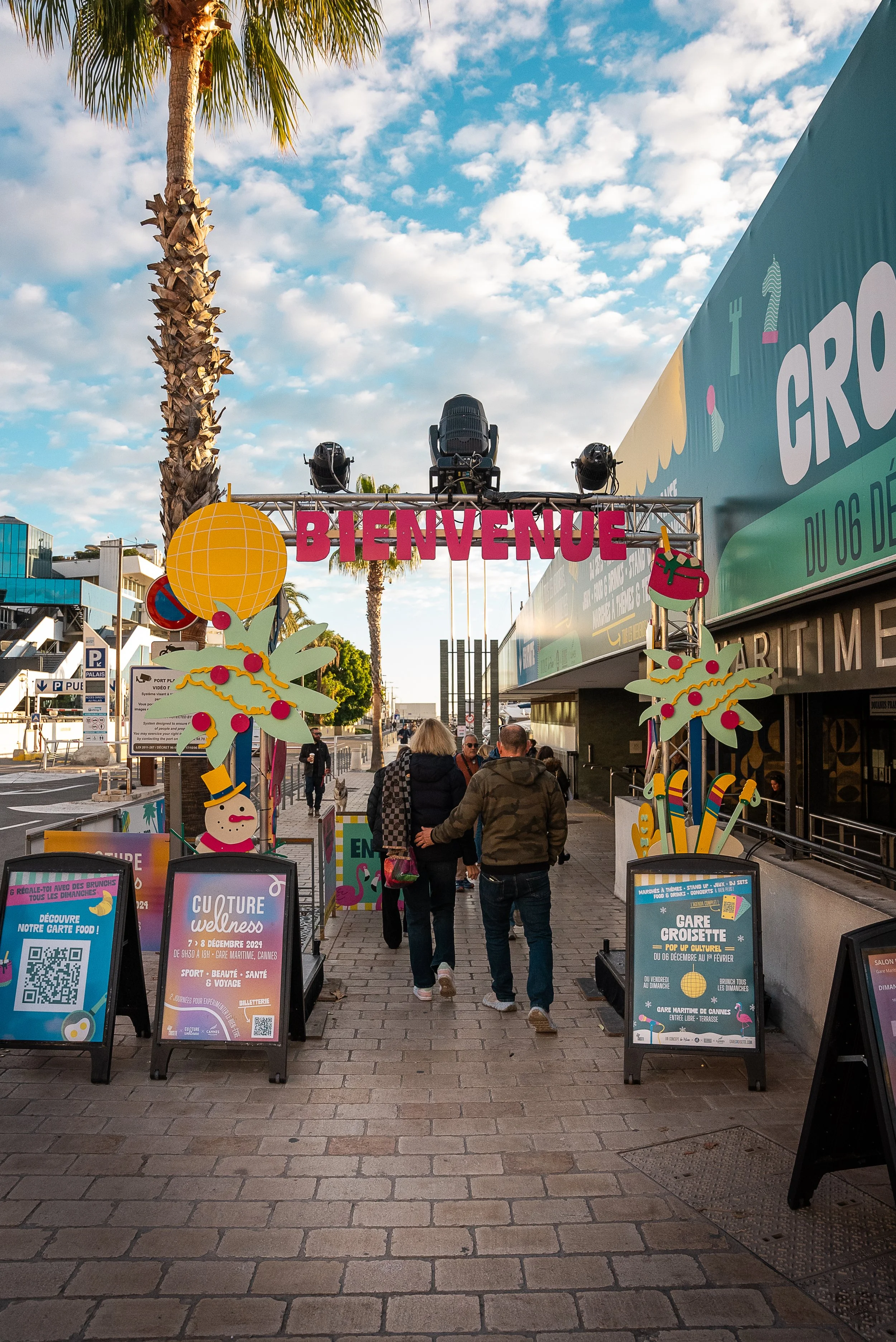 Entrée d'un événement avec un panneau 'BIENVENUE', décorations de Noël, des personnes marchant, en plein jour avec un ciel nuageux et des palmiers.