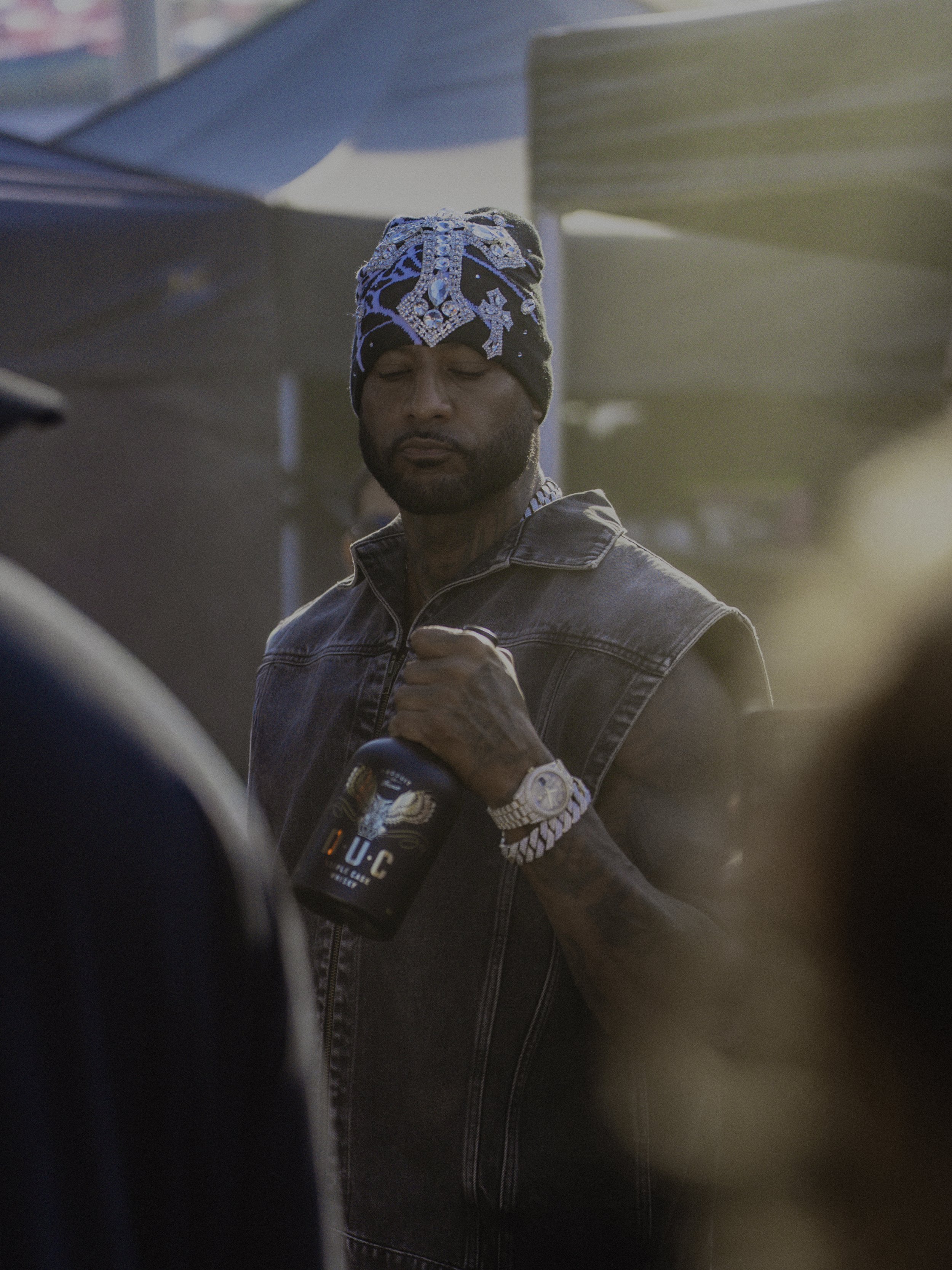 Un homme portant un bonnet coloré, une veste en denim, tenant un gobelet et portant une montre en or, dans un environnement extérieur