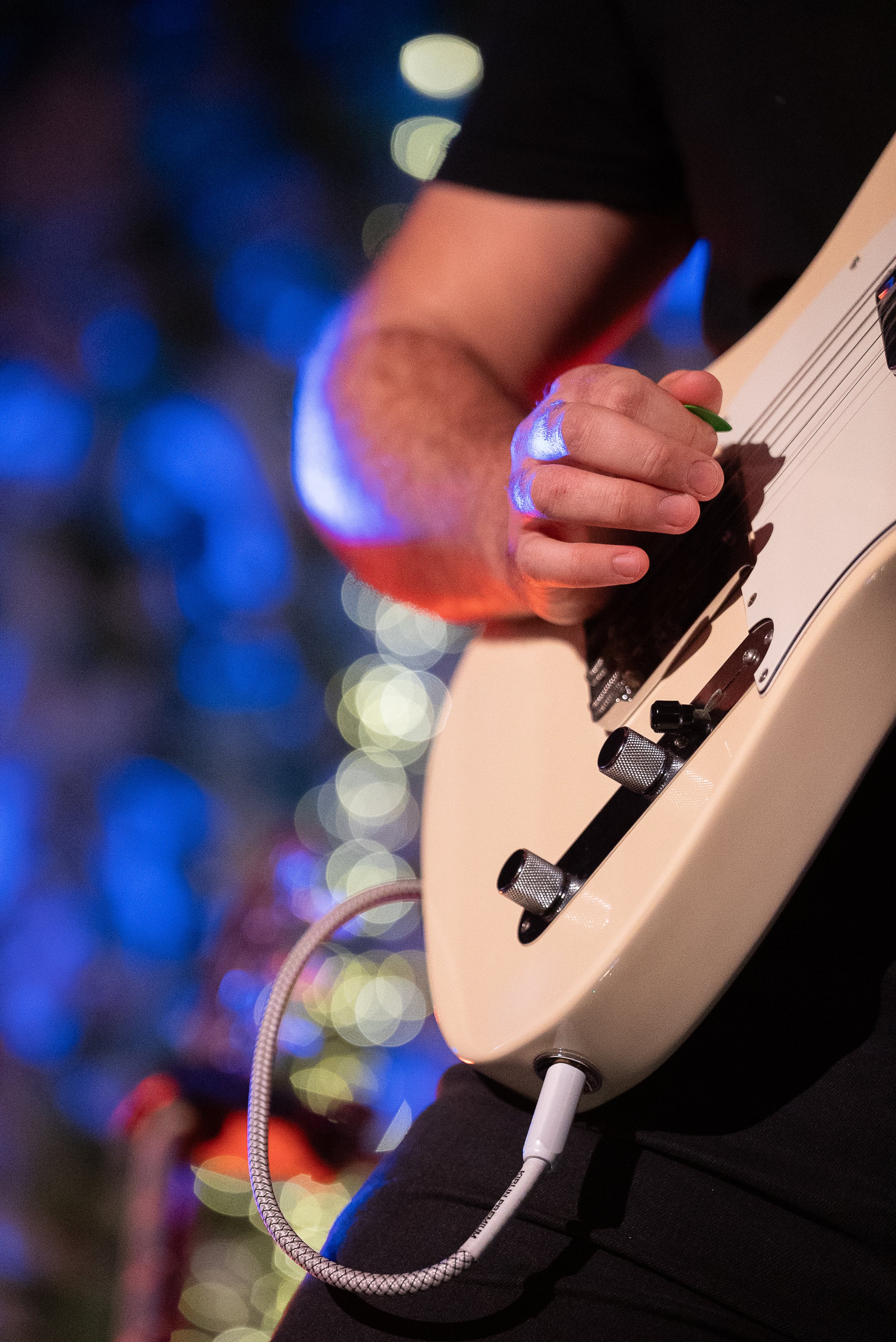 Main d'un guitariste jouant d'une guitare électrique, avec un fond flou de lumières colorées.