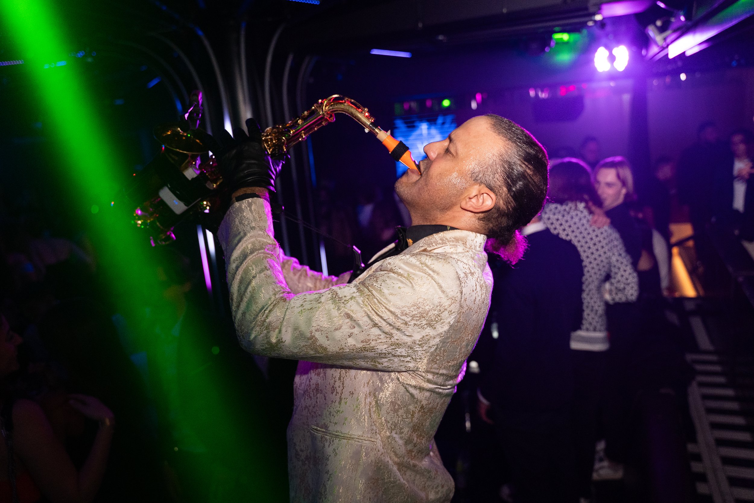 Un homme habillé d'un tuxedo beige joue du saxophoniste lors d'une soirée dans un club, avec des personnes dansant et des lumières colorées en arrière-plan.