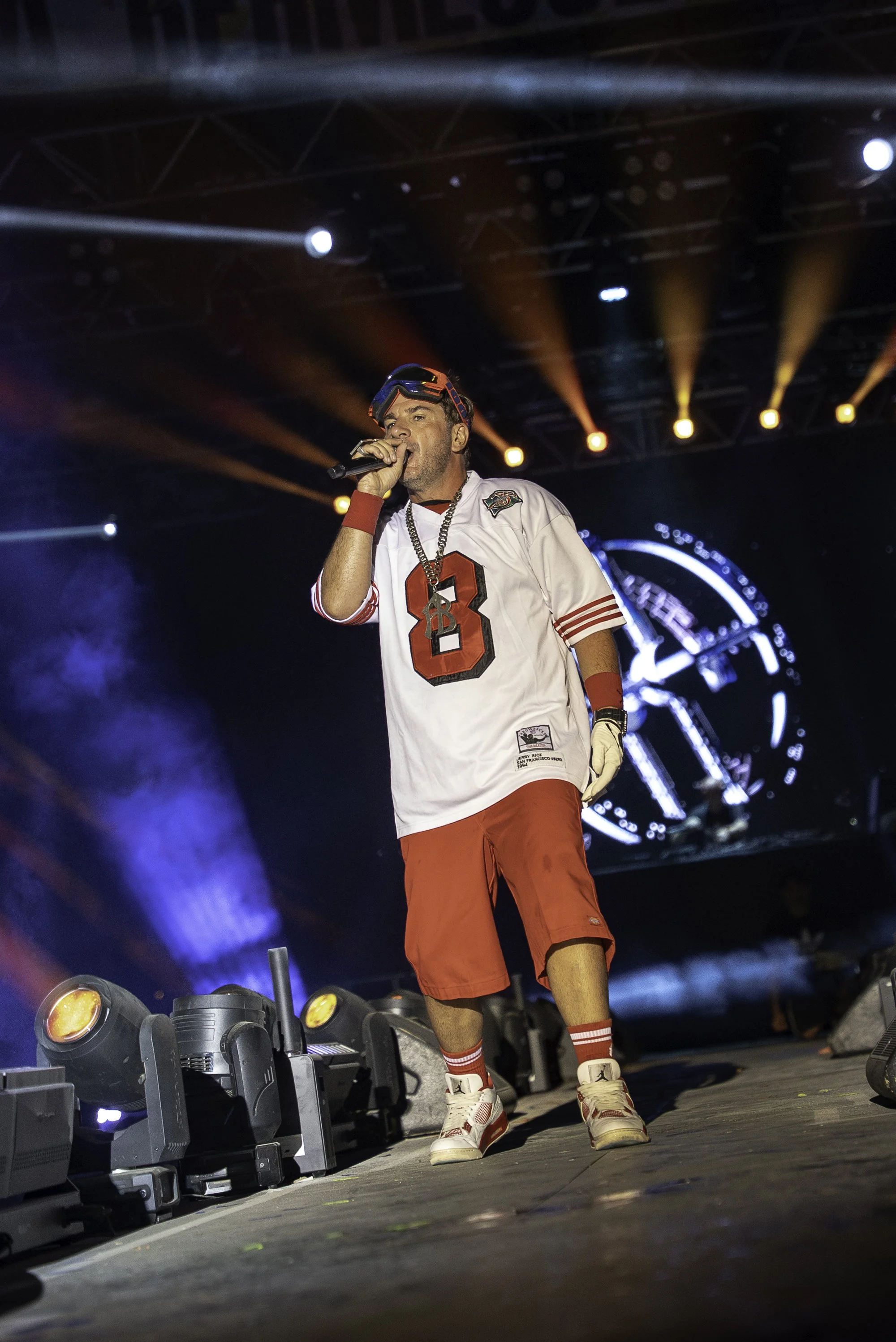 Un homme en vêtements de sport, notamment un maillot de football avec le numéro 3, un casque de ski, et une chaîne autour du cou, chante sur scène avec des lumières colorées et un écran en arrière-plan.