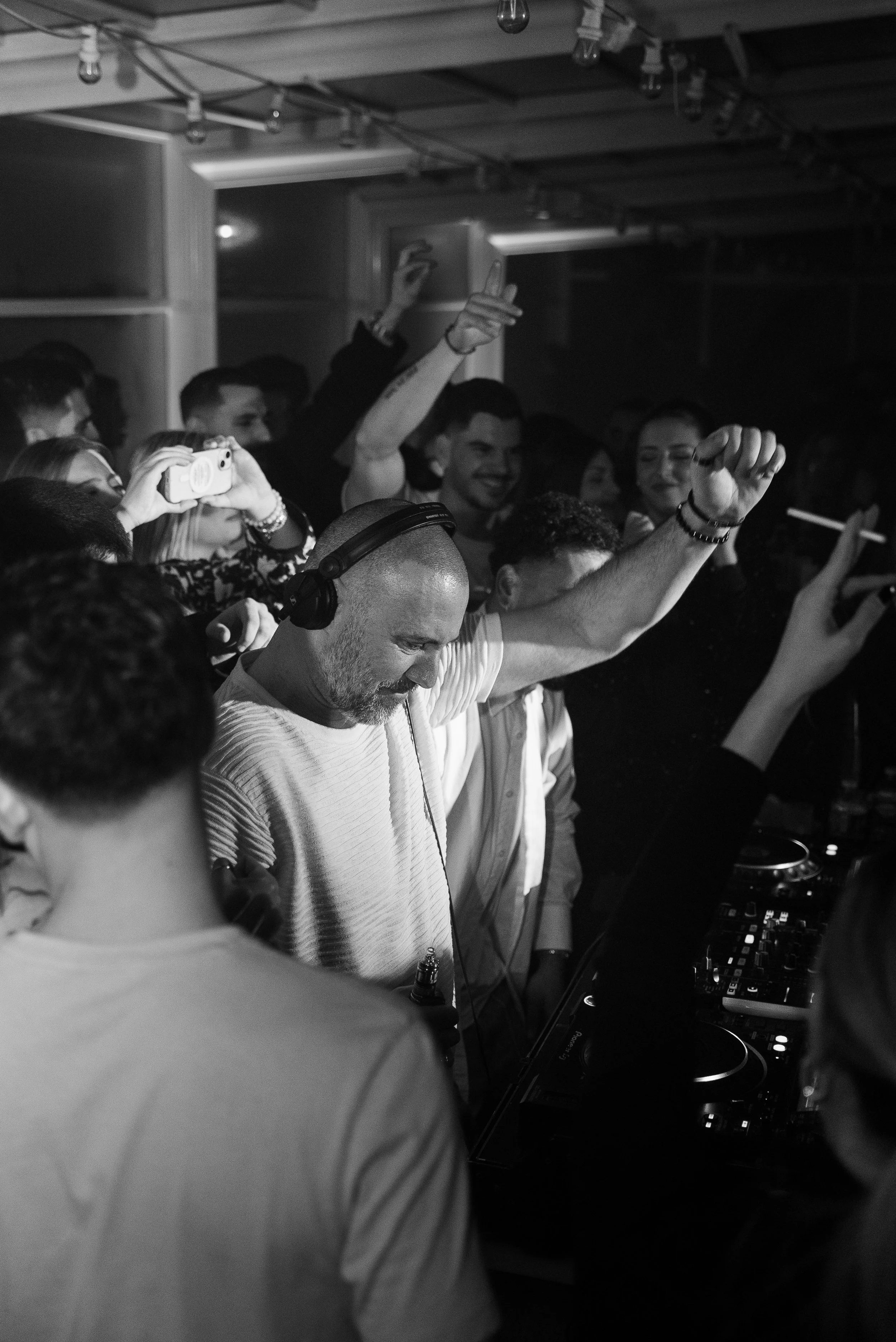 Un DJ portant des écouteurs au premier plan dans une foule qui danse et profite de la musique, en noir et blanc.