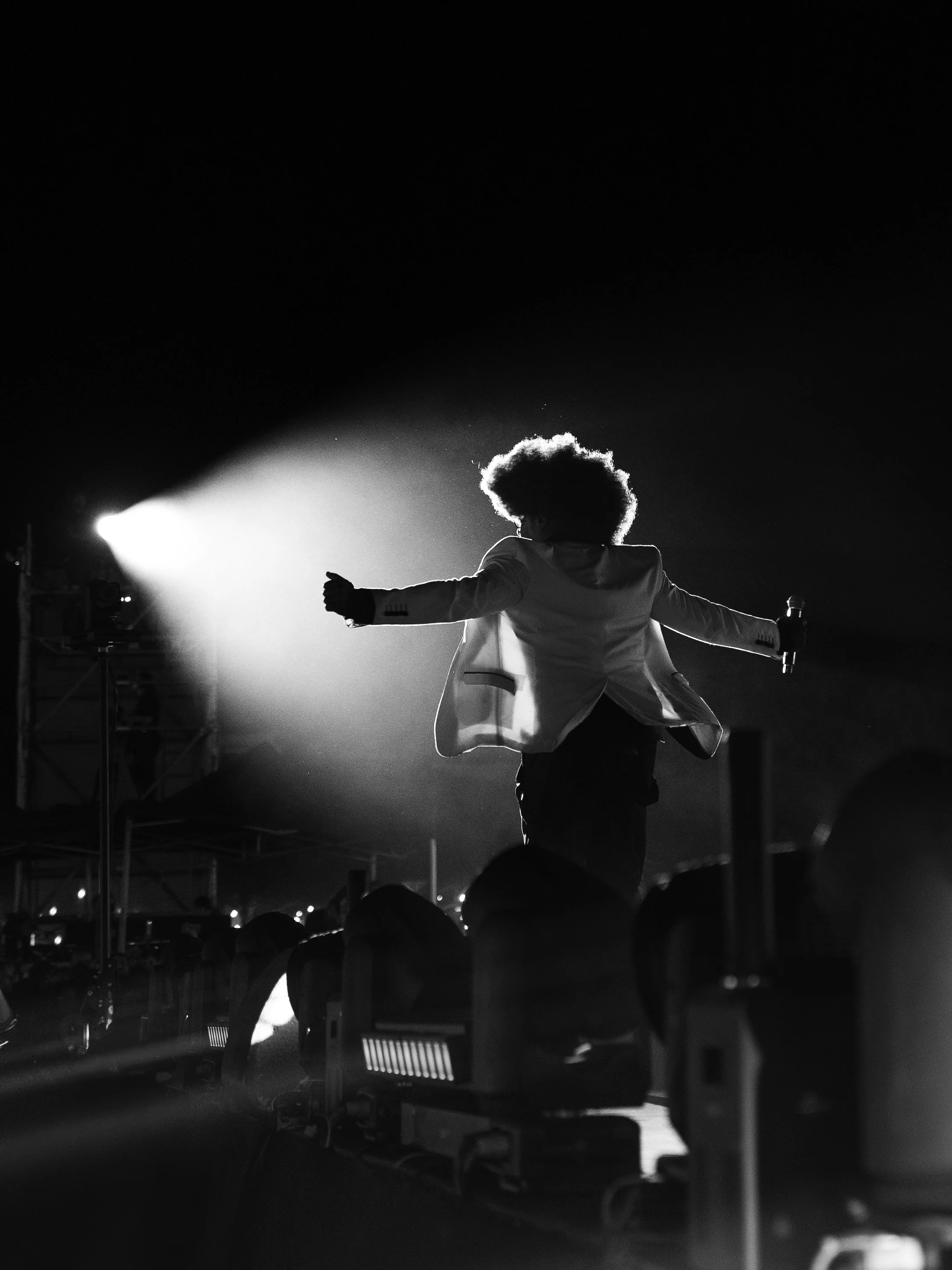 Une personne avec des cheveux volumineux sur scène, vue de dos, tenant un microphone dans chaque main, illuminée par un projecteur bright dans une ambiance de concert en noir et blanc.