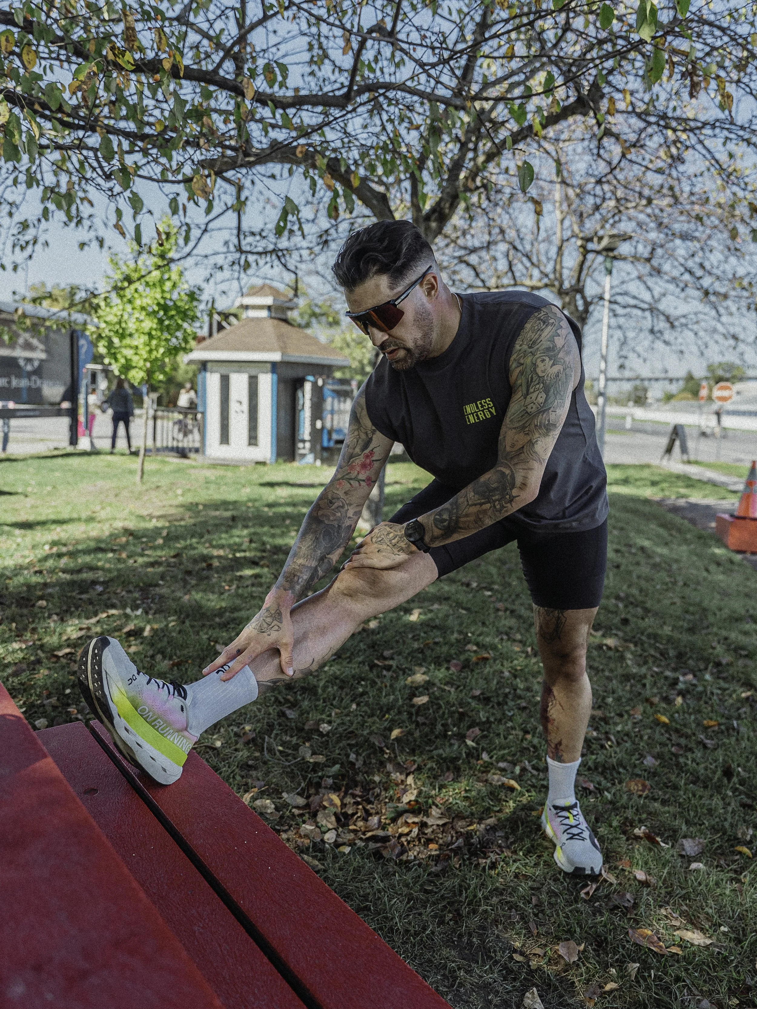 Homme faisant des étirements en extérieur sous un arbre en automne, portant un t-shirt sans manches, des shorts, des baskets et des lunettes de soleil.