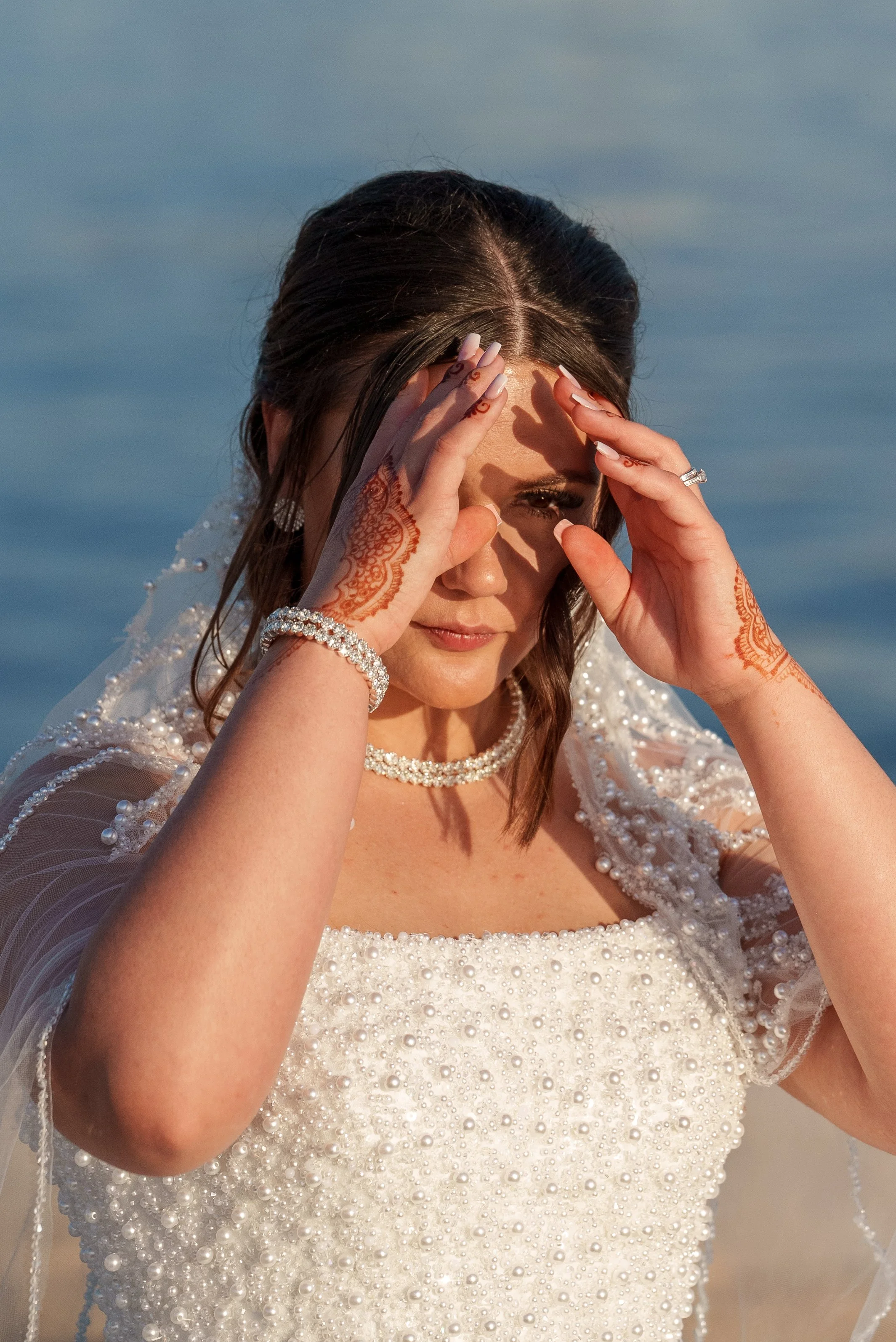 Une femme en robe de mariée avec des perles, se tenant près de l'eau, avec les mains sur le front, prêtant attention à quelque chose.