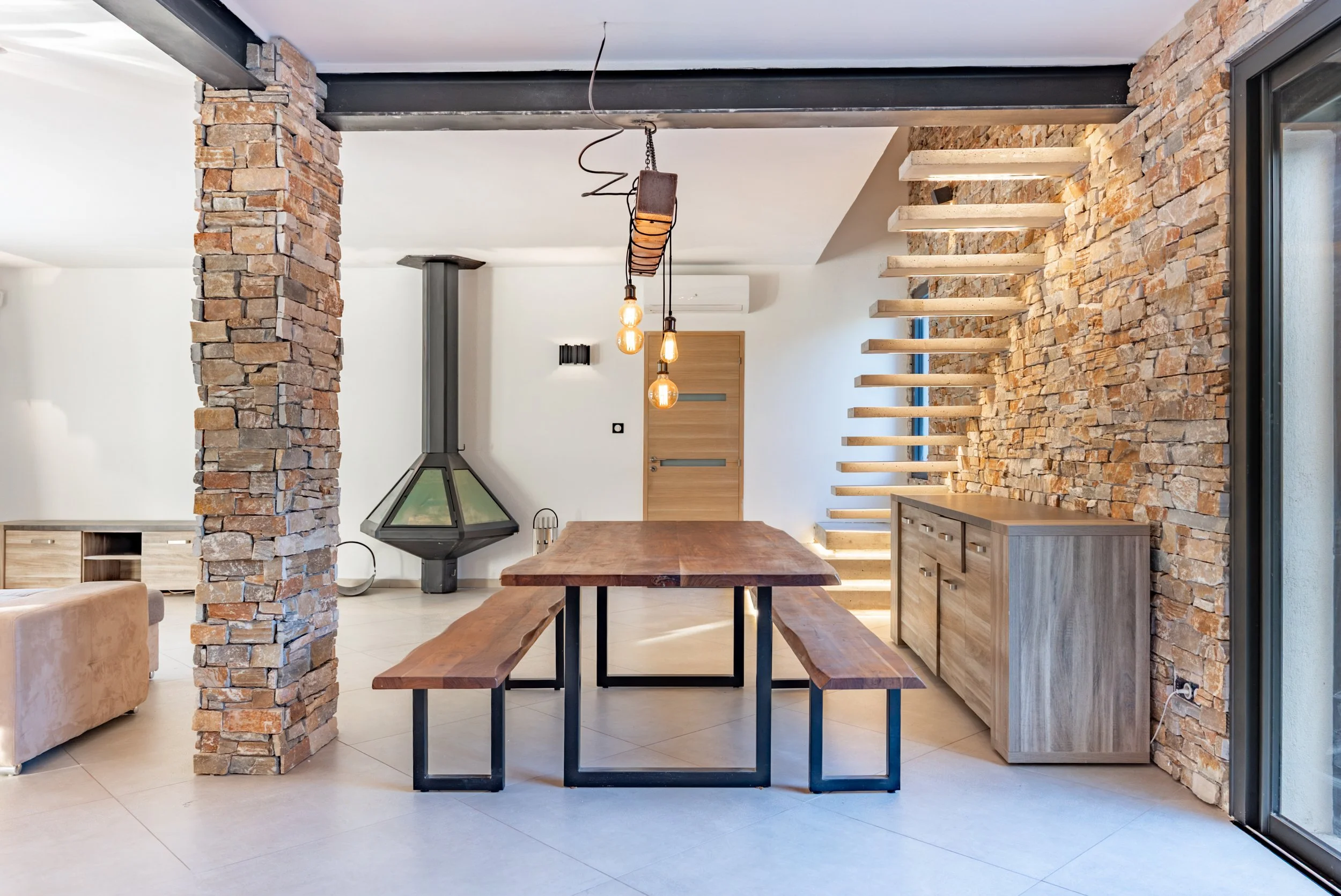 Intérieur d'une maison avec murs en pierres, une table en bois avec bancs, cheminée en forme de cône, escalier en bois flottant, meuble en bois et une porte en bois moderne.