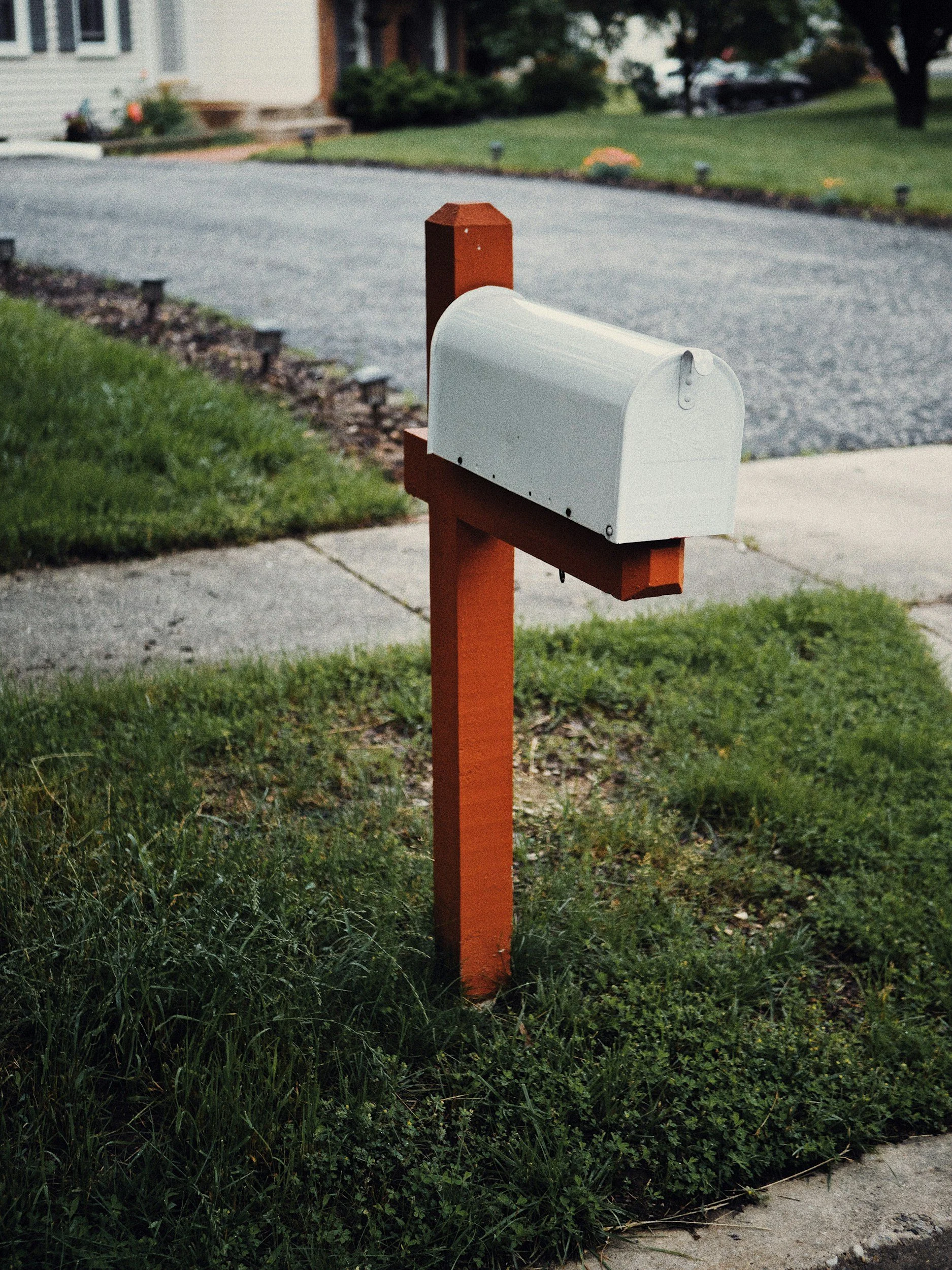 A white mailbox on an orange post situated on a lawn next to a sidewalk, with a residential street and houses in the background.