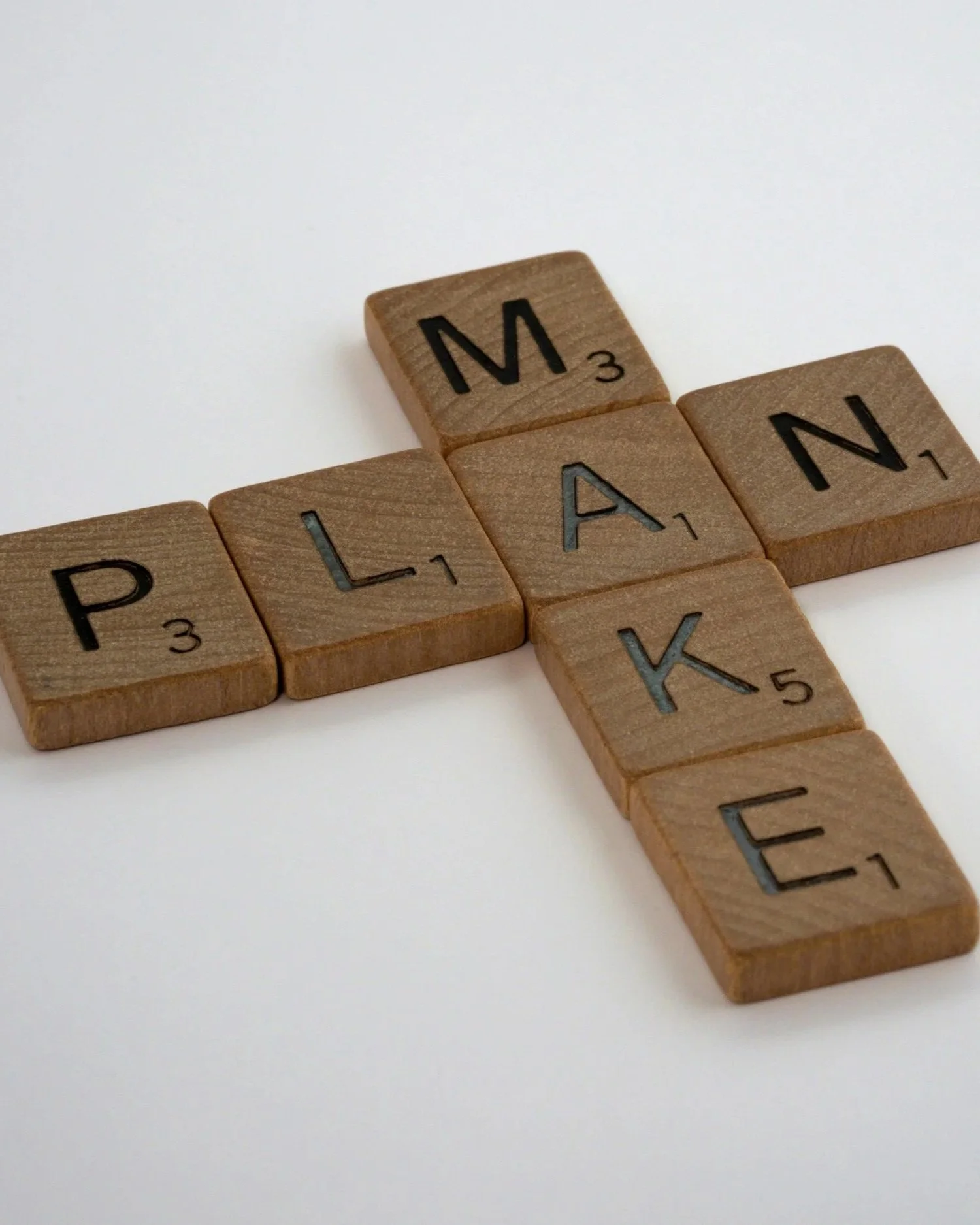 A close-up of wooden Scrabble tiles arranged on a white surface, forming the words 'MAKE' and 'PLAN.