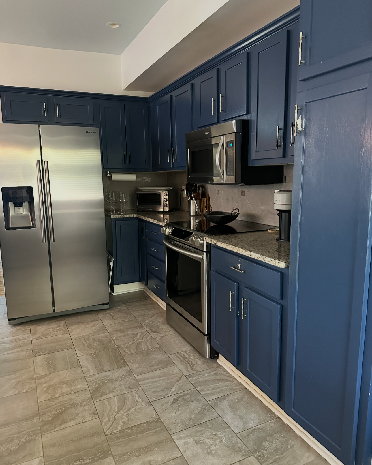 Kitchen with blue cabinets, stainless steel refrigerator, stove, microwave, toaster oven, granite countertops, tiled floor, and various kitchenware.