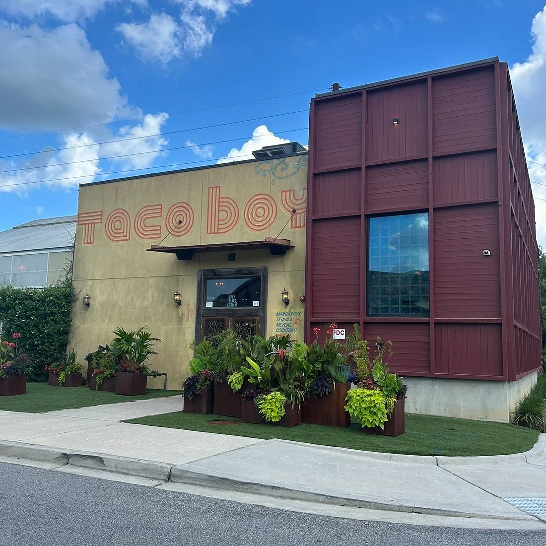Exterior of a restaurant with a tan wall and a red wooden addition, large plants and flowers in planters outside, blue sky with clouds overhead.