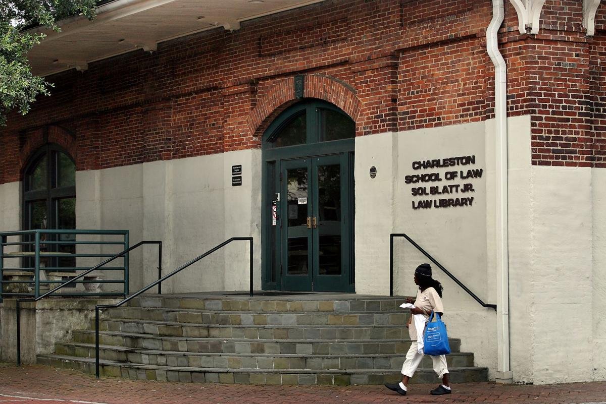 The entrance to the Charleston School of Law, Sol Blatt Jr. Law Library, featuring a brick building with steps leading up to a green double door, a woman walking by holding papers and a blue bag.