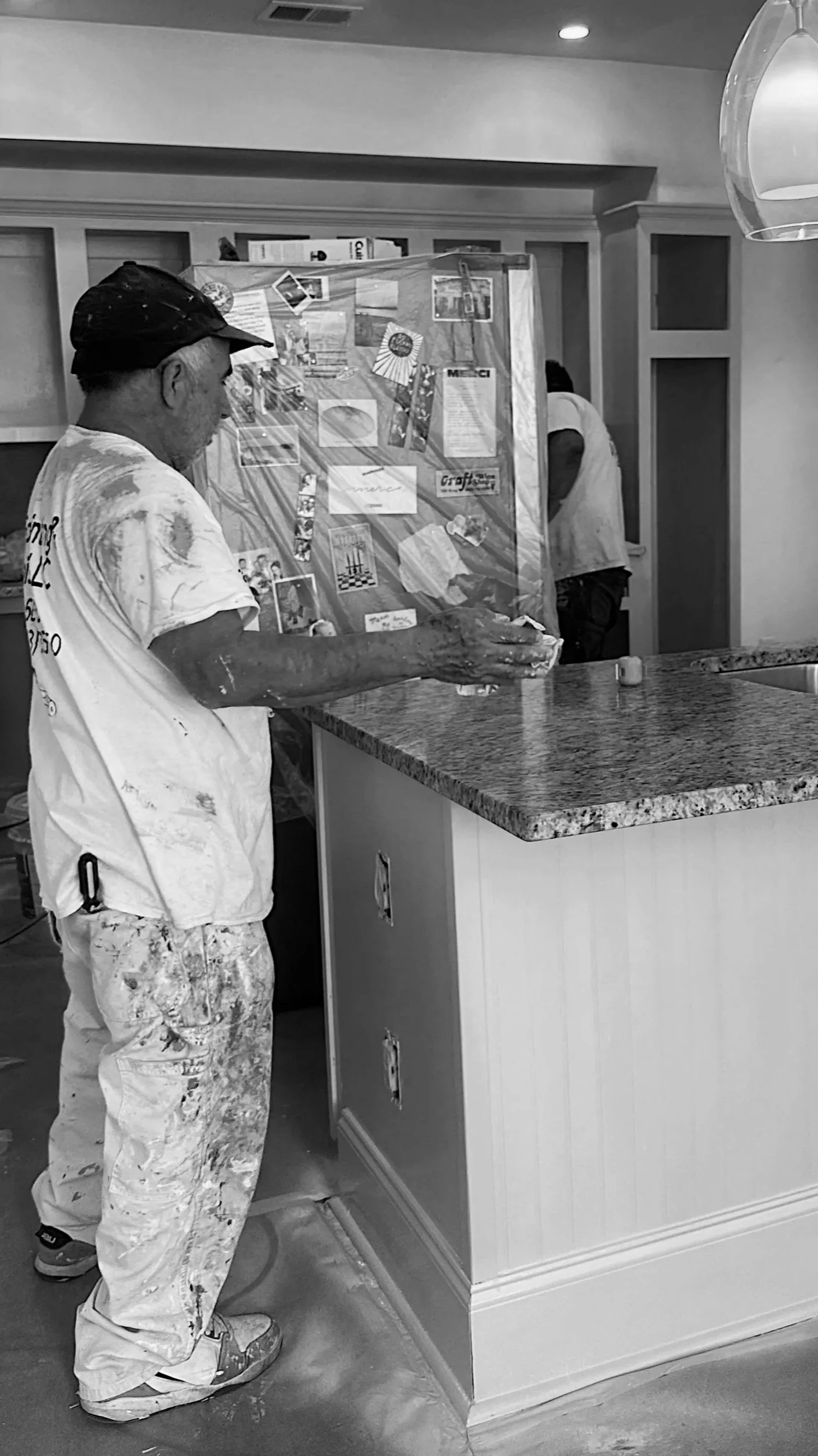 A man (Octavio) wearing painter's clothes, with paint stains, is holding a clear plastic sheet with various pictures and cards attached to it. He is standing in a kitchen under renovation, with a granite countertop and a white cabinet.