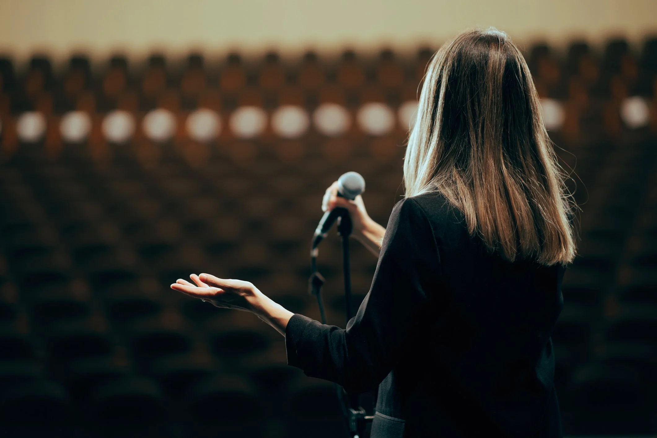 A woman with long blonde hair in a black blazer holds a microphone on a stage in an empty auditorium, facing away from the camera.