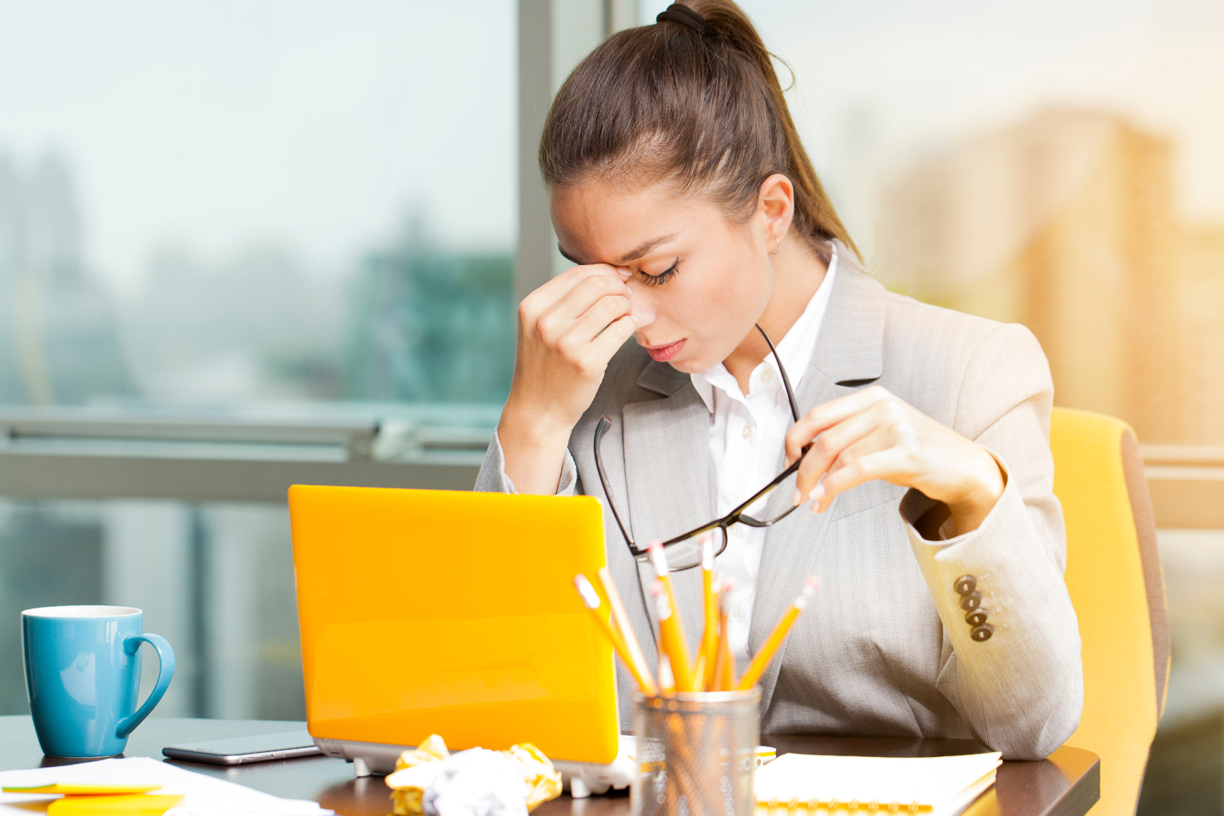 A stressed woman in a gray business suit sitting at a cluttered desk, pinching the bridge of her nose, holding her glasses, with a yellow tablet, a blue mug, and scattered papers and pens in front of her.
