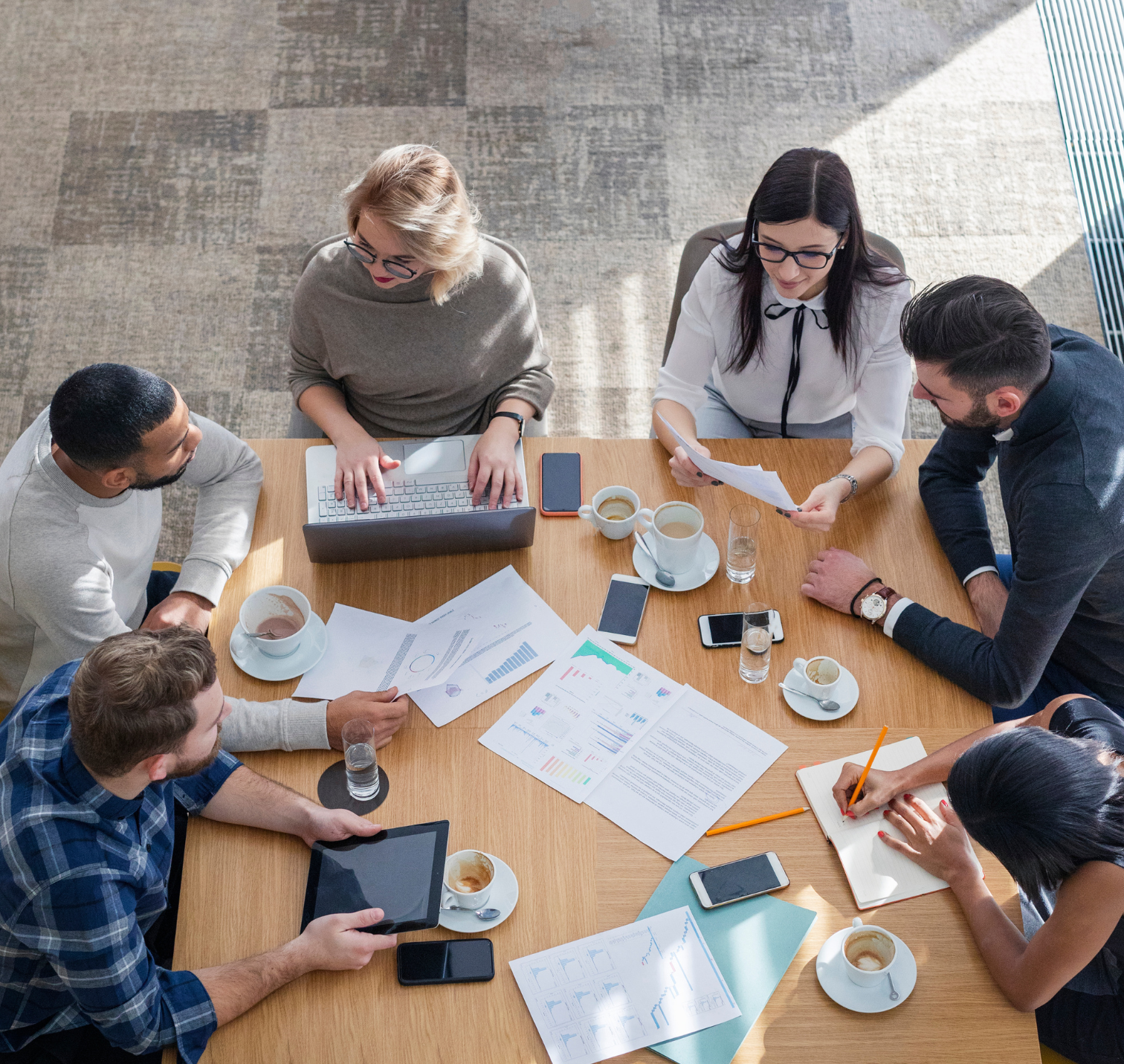 Overhead view of a diverse group of eight people gathered around a wooden conference table with laptops, tablets, papers, notebooks, smartphones, cups of coffee, glasses of water, and pens, engaged in a business meeting.