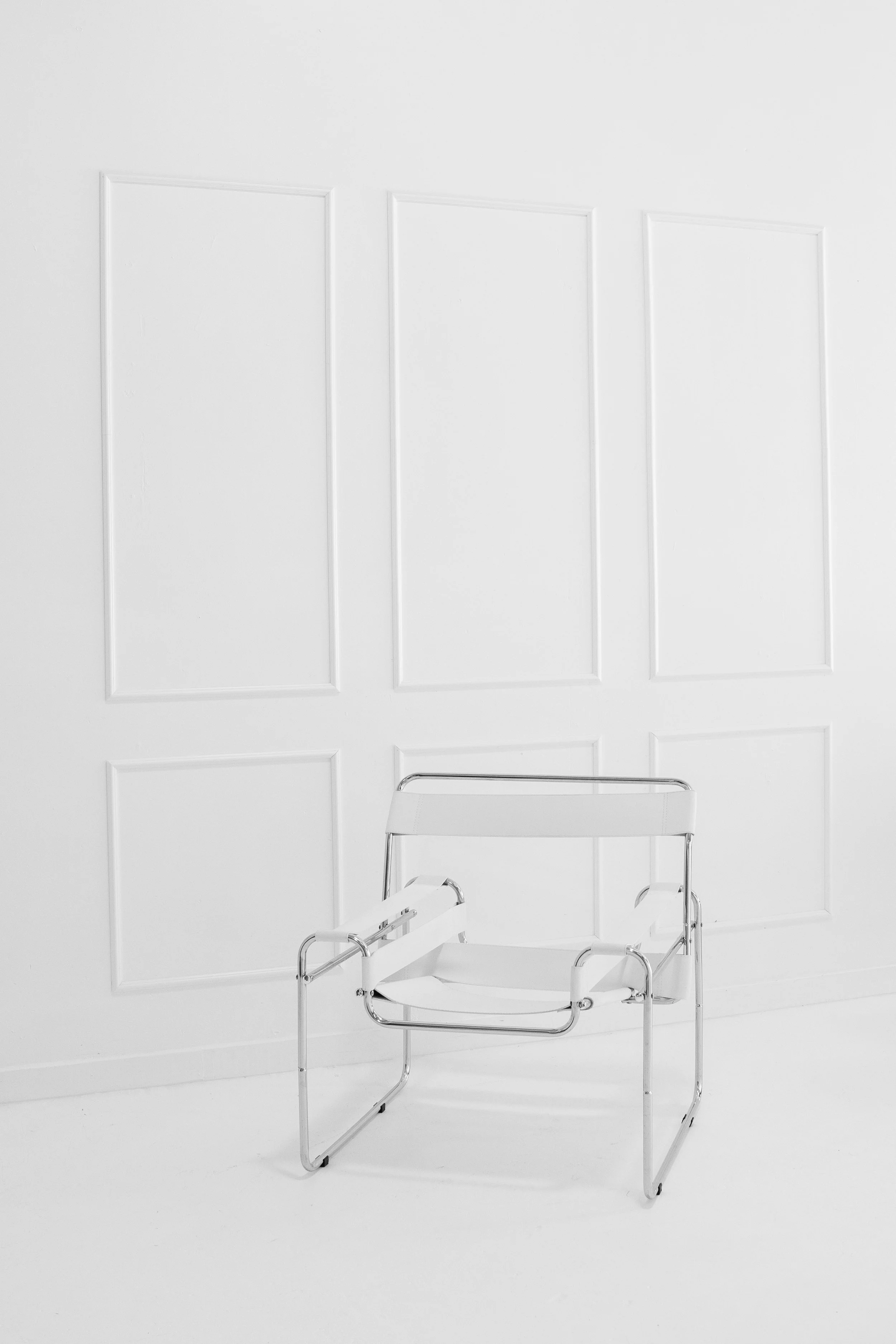 A white chair with chrome frame positioned against a white wall with panel molding in a minimalistic setting.