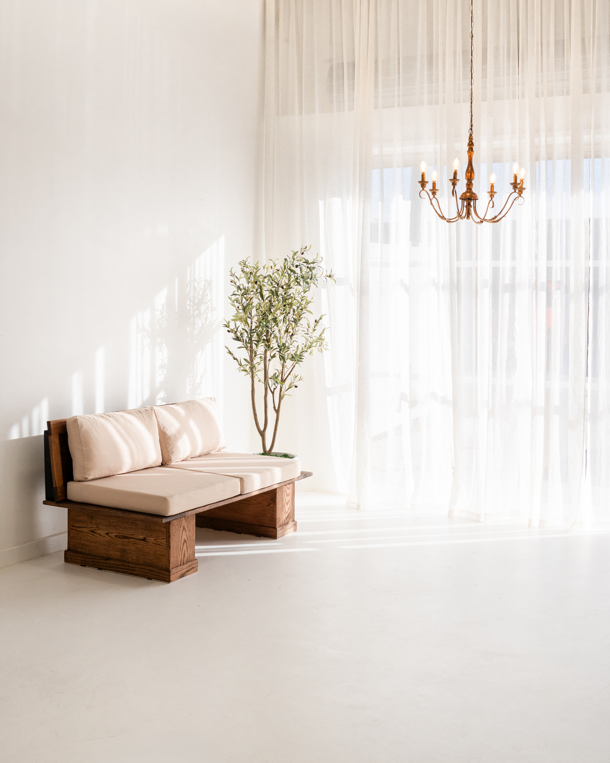 Minimalist living room with a wooden bench, beige cushions, a potted plant, sheer curtains, and a chandelier.
