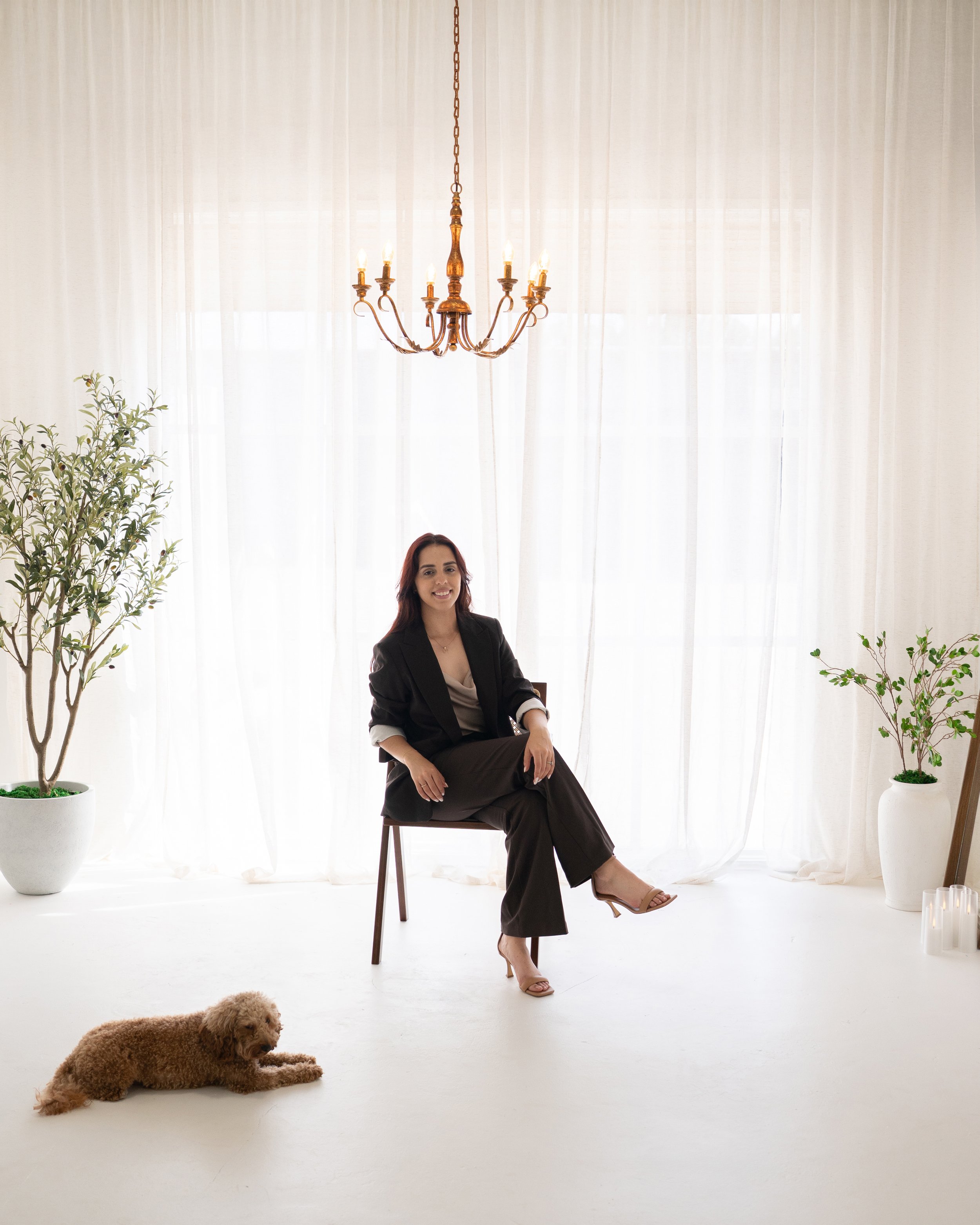 A woman in a black blazer and brown pants sitting on a chair, smiling, with a small dog lying on the floor beside her. The background features white curtains, a chandelier, potted plants, and candles.