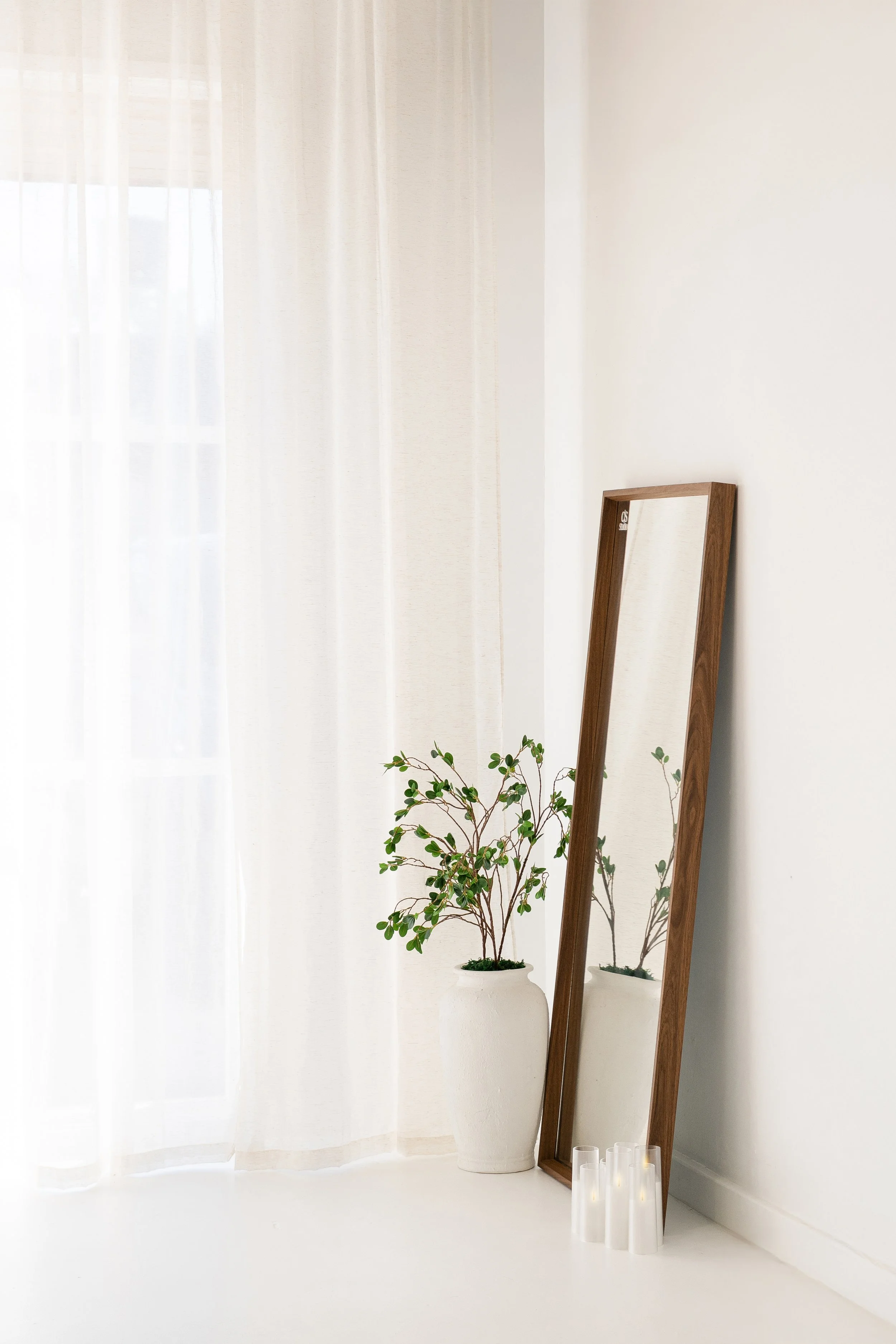 A tall wooden-framed mirror leaning against a white wall next to a potted green plant, with three white candles on the floor nearby, and light-colored curtains covering a window.