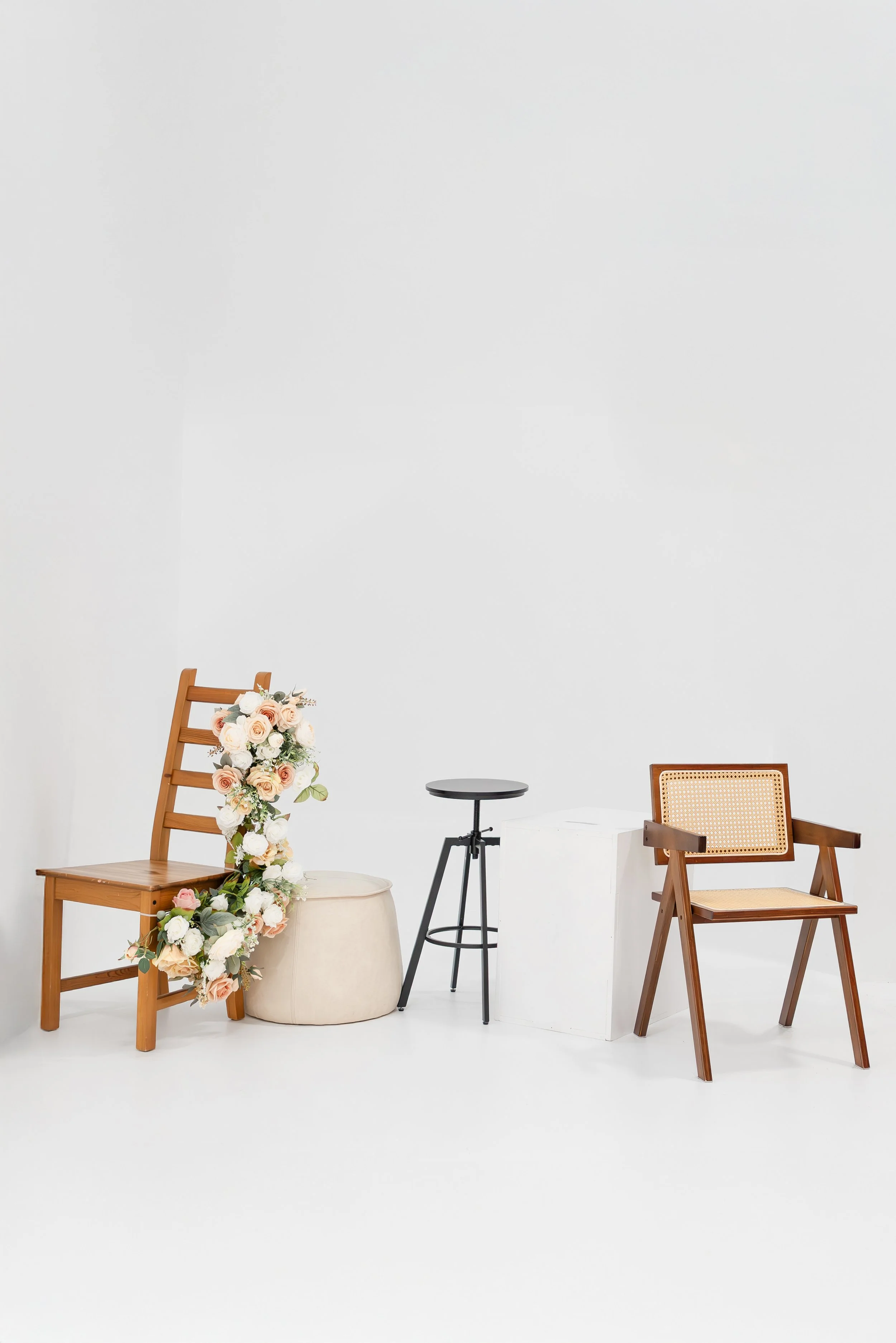 Minimalist arrangement of two wooden chairs, a small black stool, and a white cube in a white room, with floral decoration on one chair.