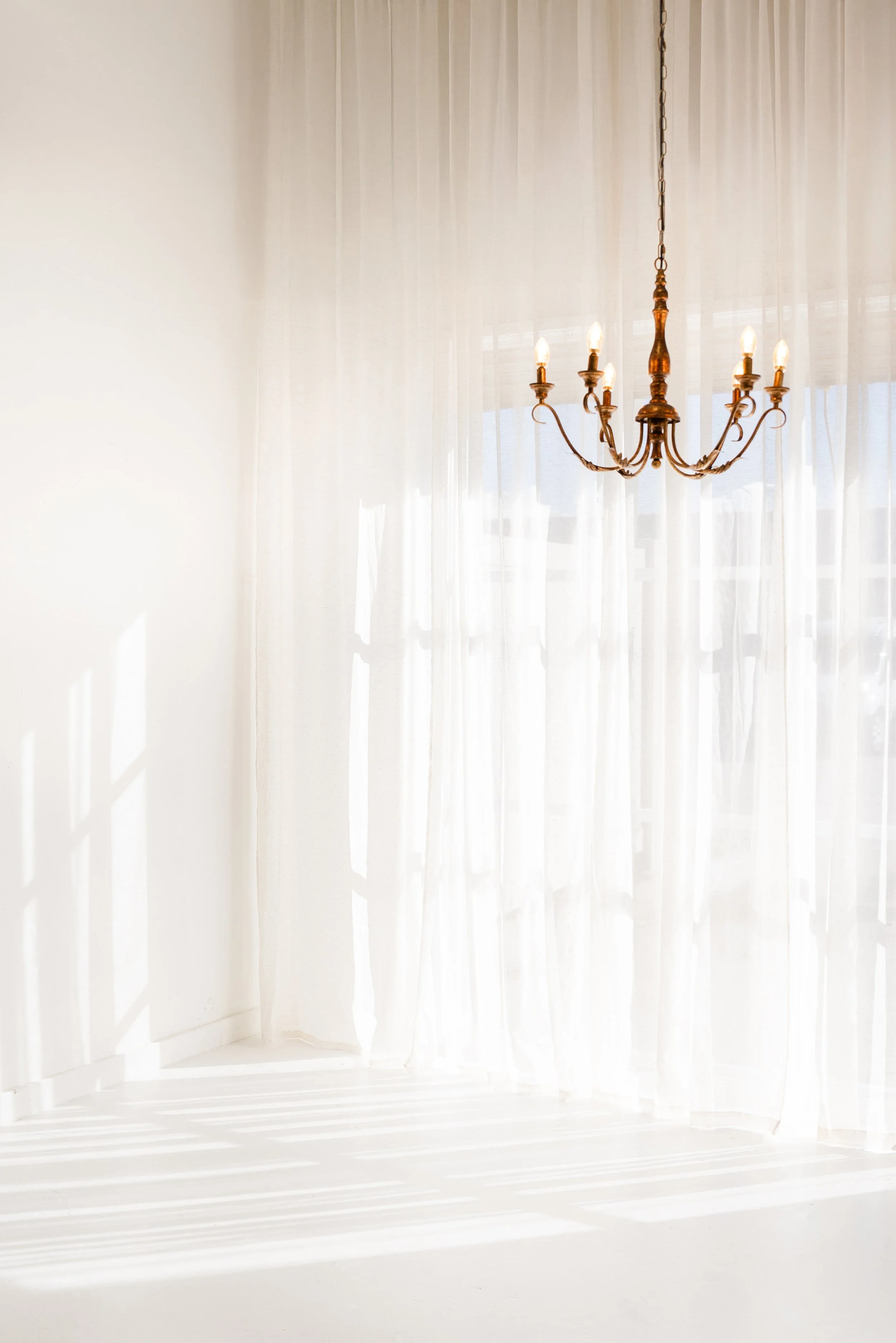 A chandelier hanging from the ceiling in front of sheer white curtains with sunlight filtering through.
