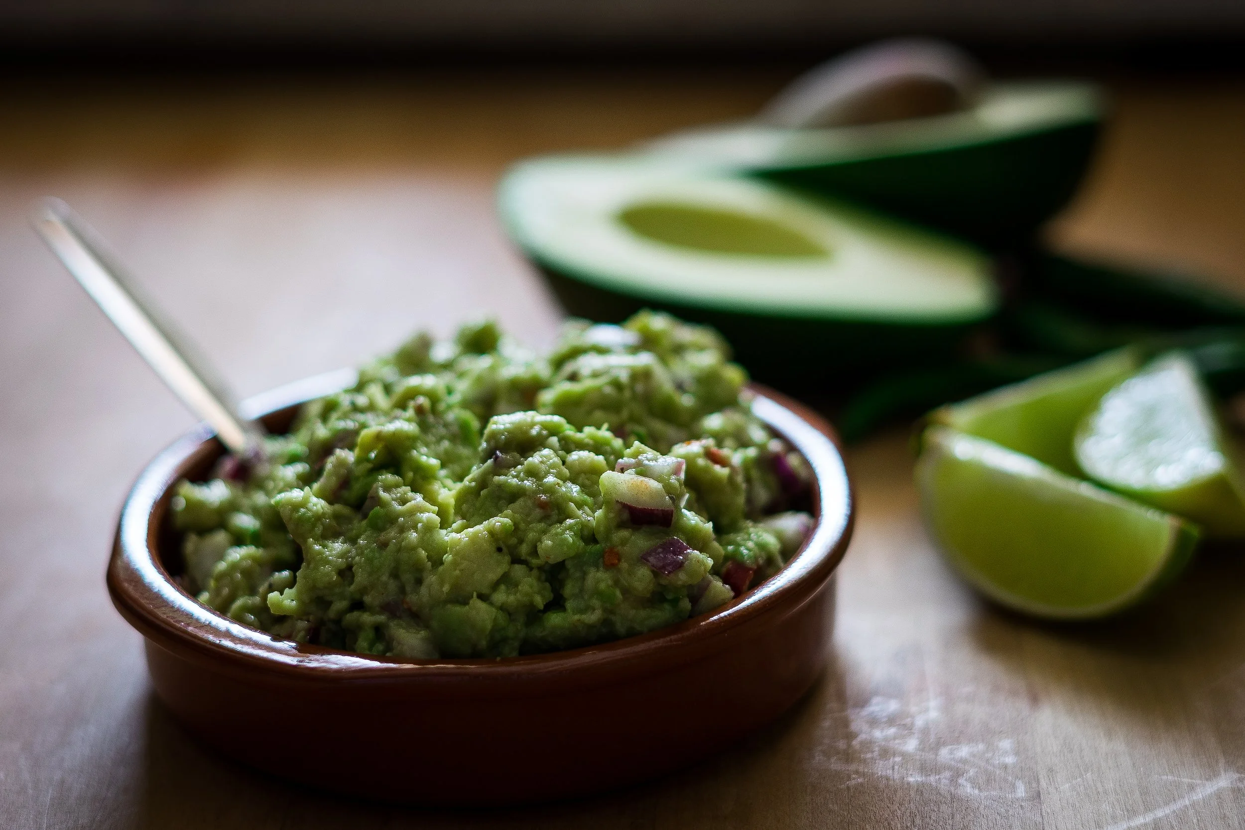 A small wooden bowl filled with mashed avocado, placed on a wooden surface with sliced avocado halves in the background.