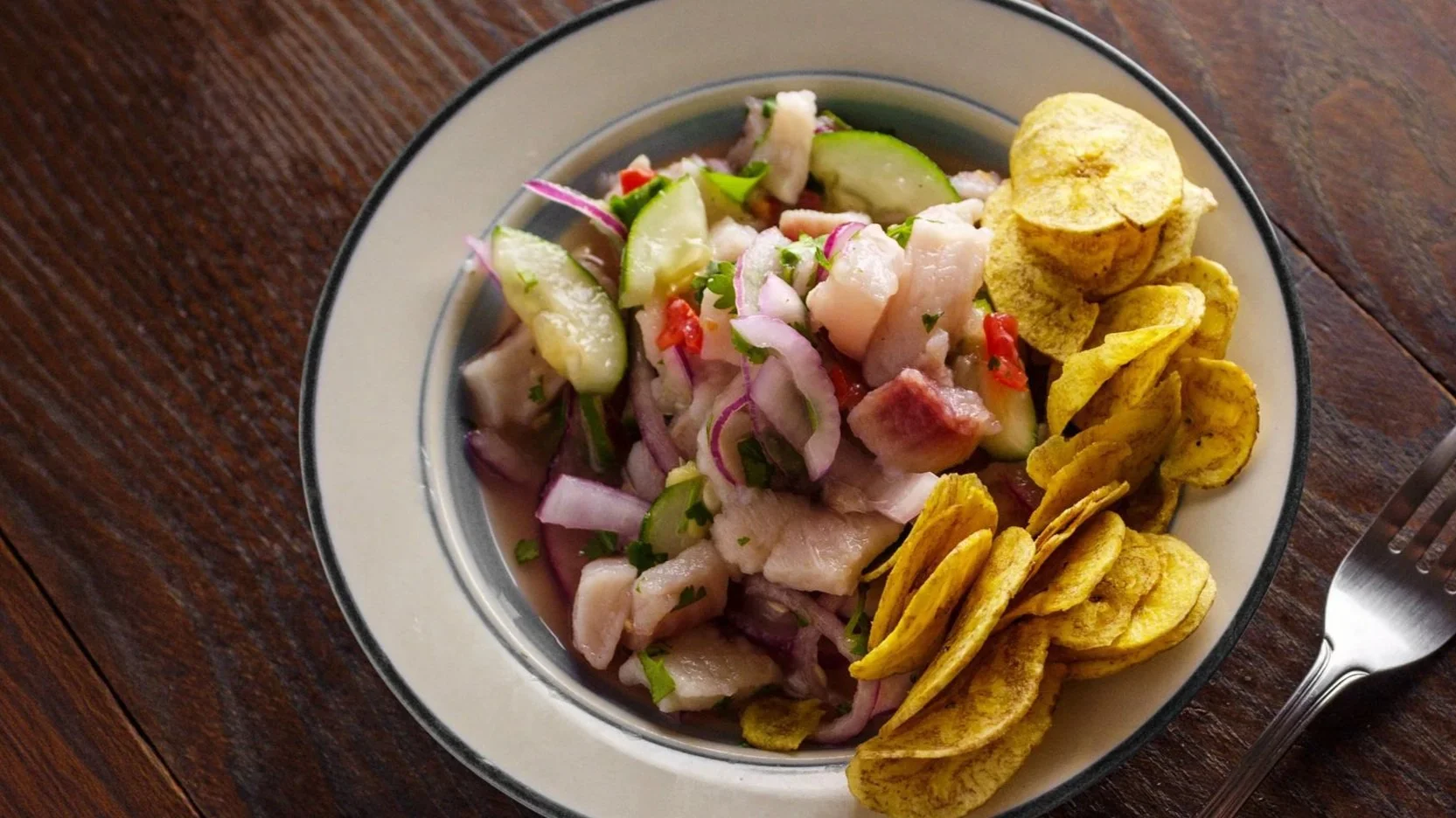 A bowl of ceviche with sliced cucumbers, red onions, fish, and red chili pieces, served with plantain chips on the side, on a wooden table.