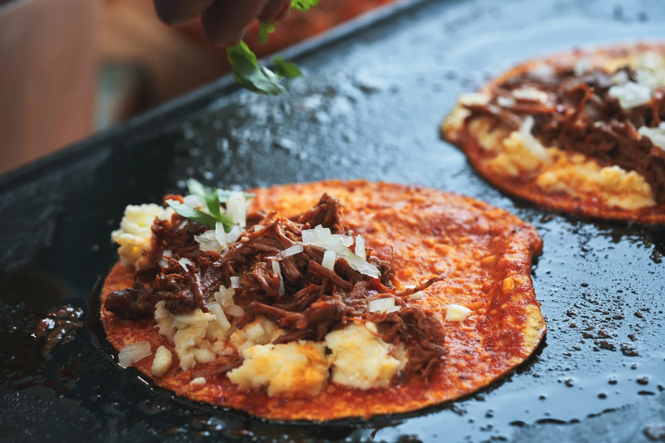 Close-up of carne adobada on tortilla being prepared on a black skillet, topped with chopped onions and cilantro.