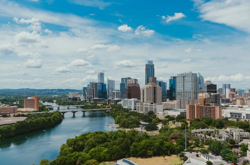Austin city skyline with tall modern skyscrapers, a river with bridges, and green trees in the foreground under a partly cloudy sky.