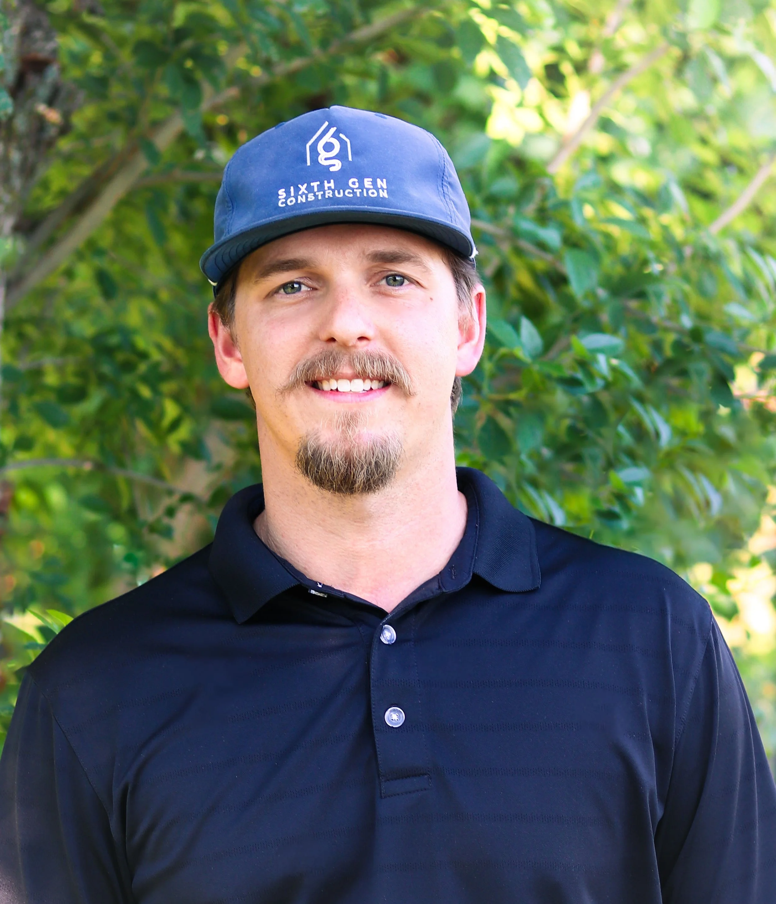 A man with blue eyes, wearing a blue cap with a logo and the words 'Sixth Gen Construction', and a black polo shirt, standing outdoors in front of green foliage.