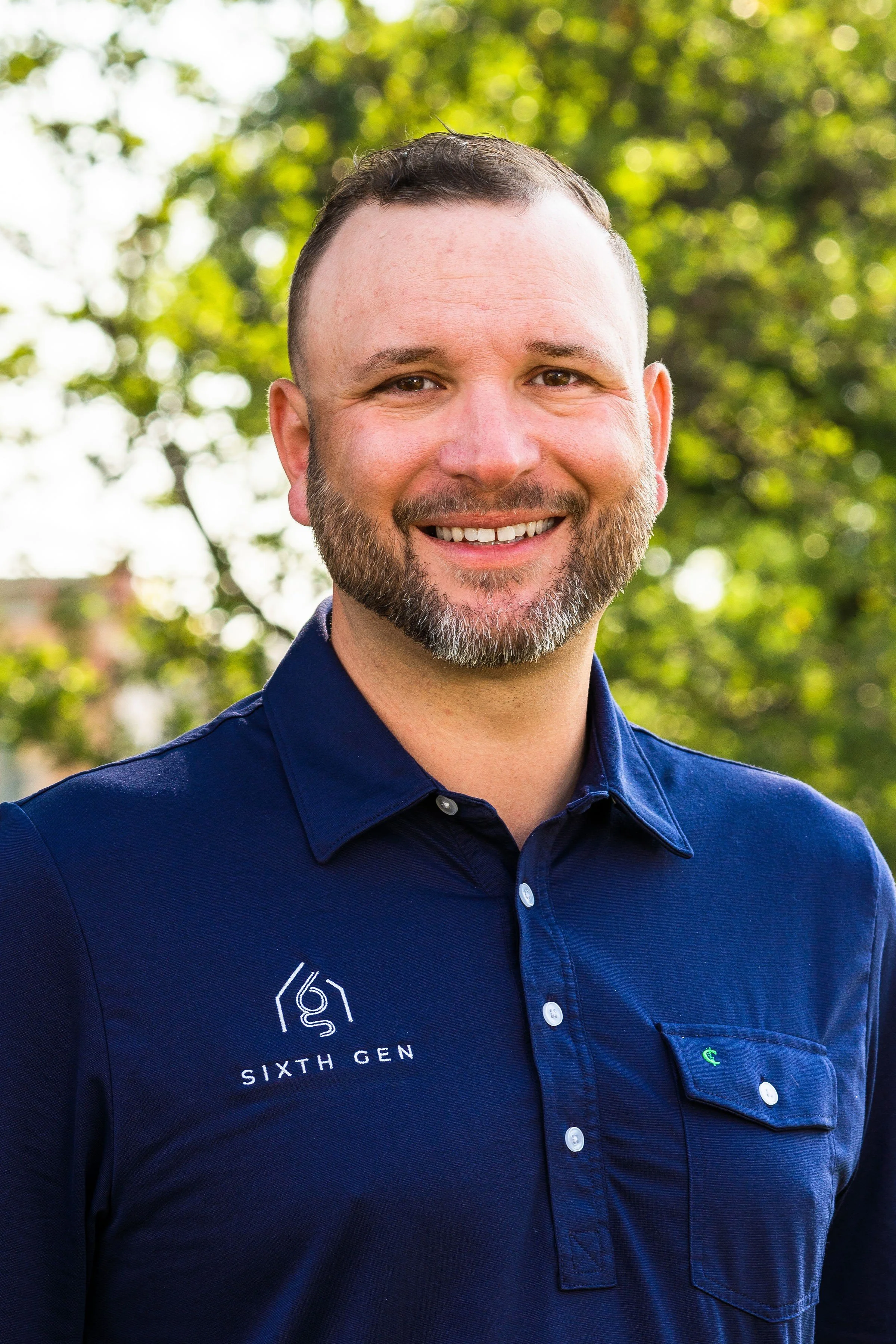 A smiling man with short brown hair and a beard, wearing a navy blue shirt with a logo that says 'Sixth Gen,' standing outdoors with blurred green trees in the background. Josh Herrington, Sixth Gen Construction CEO.