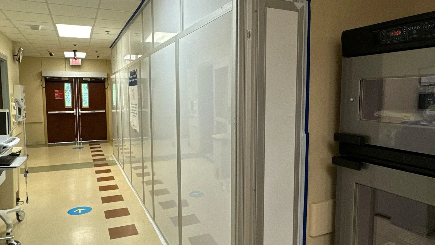Hospital corridor with a glass partition on the right, medical equipment on the left, and double doors at the end. Blue directional arrows on the floor.