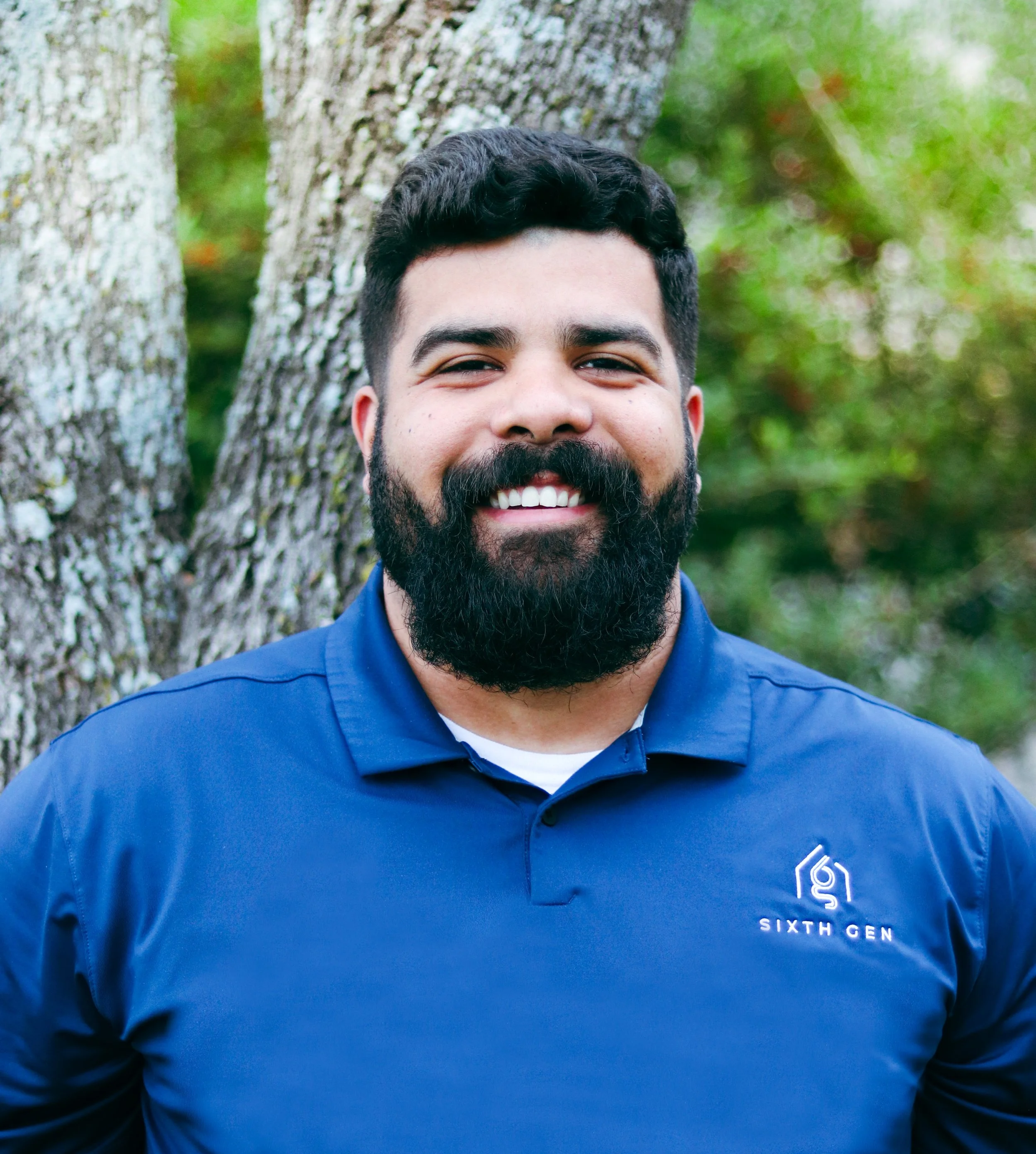A smiling man with dark hair and a full beard poses outdoors in front of a tree, wearing a blue collared shirt with a logo that reads 'Sixth Gen'.