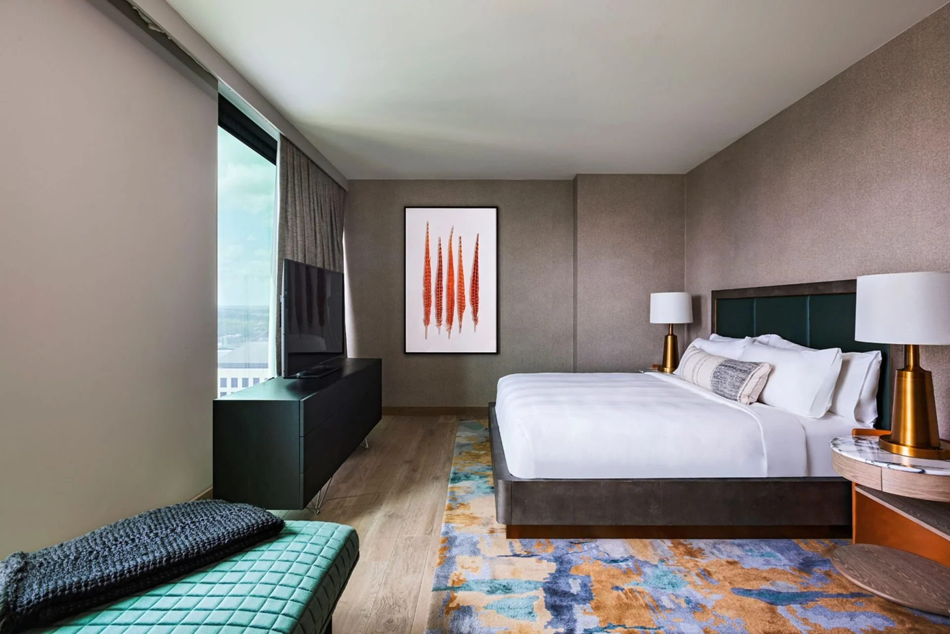 Hotel room with a large bed, side tables with lamps, a television, a bench, a painting, and a window with curtains.