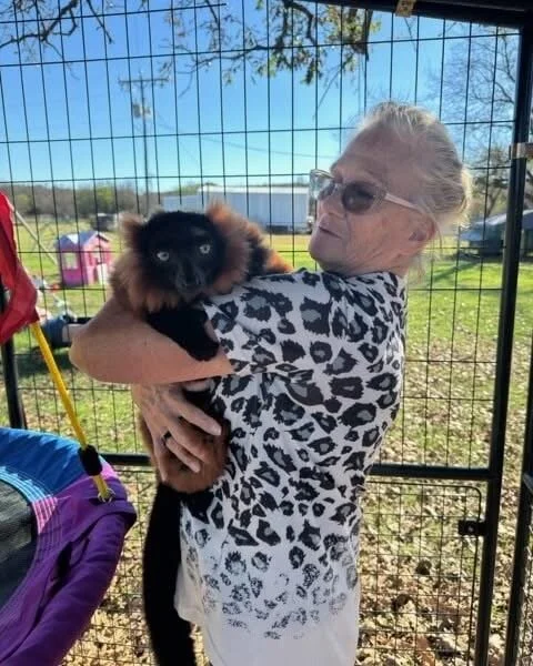 An elderly woman with glasses holding a black and brown puppy inside a fenced outdoor area.