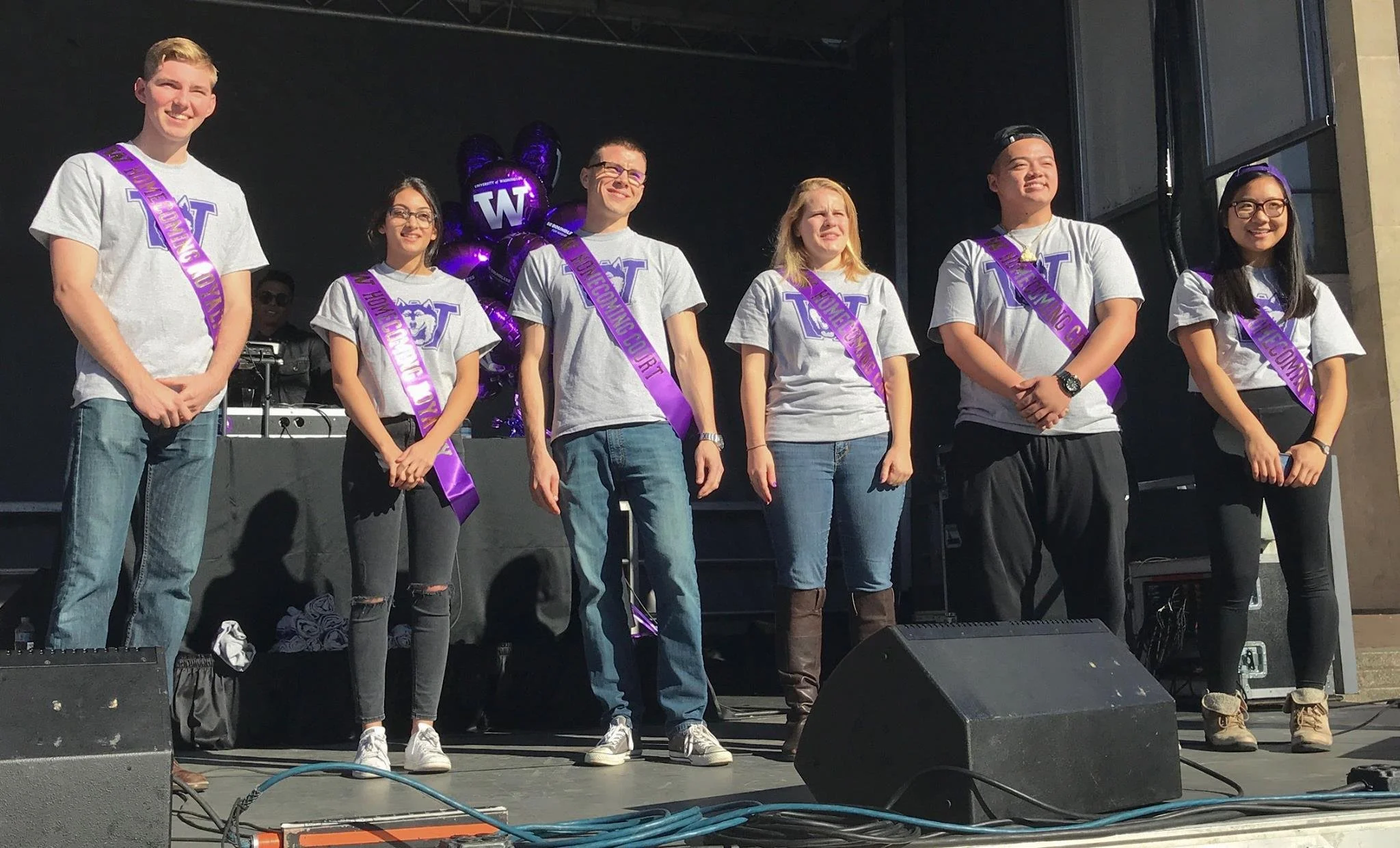 Six students standing on stage wearing gray T-shirts with a purple and white logo, each with a purple sash that reads 'Valedictorian,' 'Salutatorian,' or similar titles. Behind them is a cluster of purple balloons with a 'W' logo and a DJ setup. The students are smiling or looking at the audience.