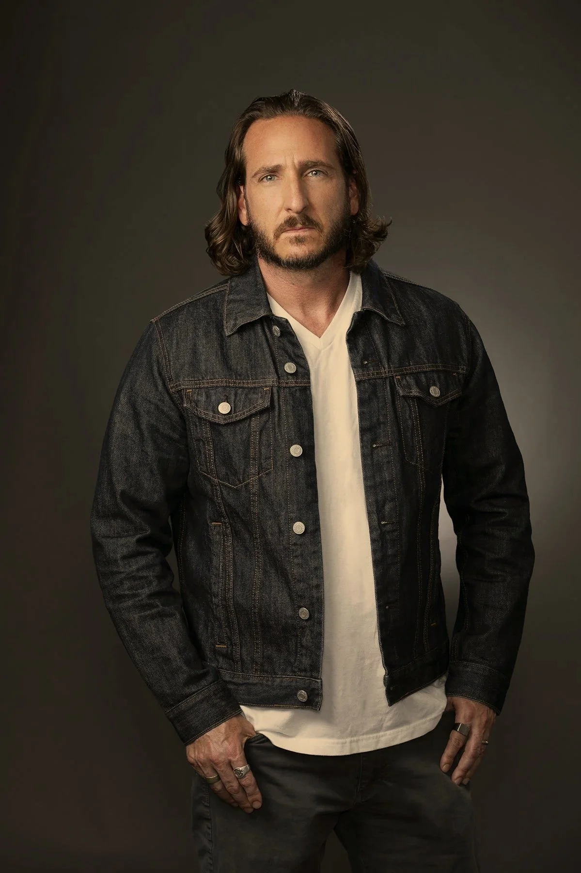 A man with shoulder-length hair and a beard, wearing a black denim jacket, white T-shirt, and dark pants, standing against a dark background.