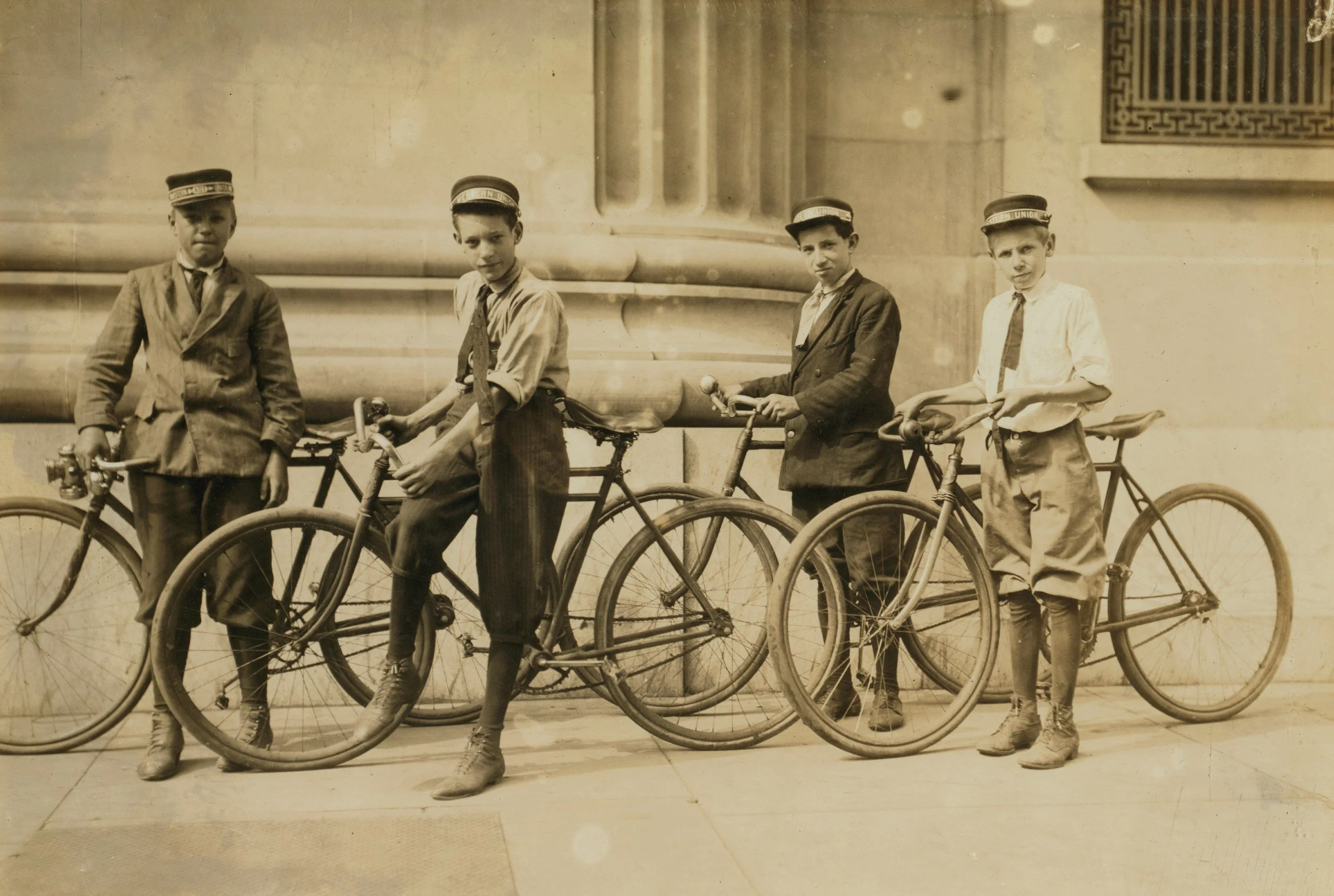 Four boys standing with bicycles in front of a building with architectural details, dressed in vintage clothing and caps.
