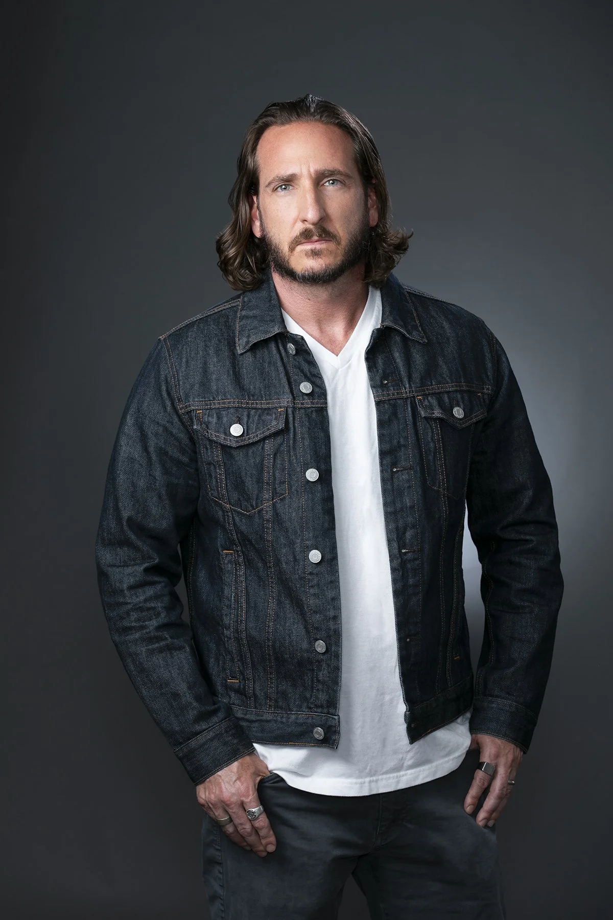 A man with shoulder-length brown hair and a beard, wearing a dark denim jacket over a white shirt, standing against a gray background.