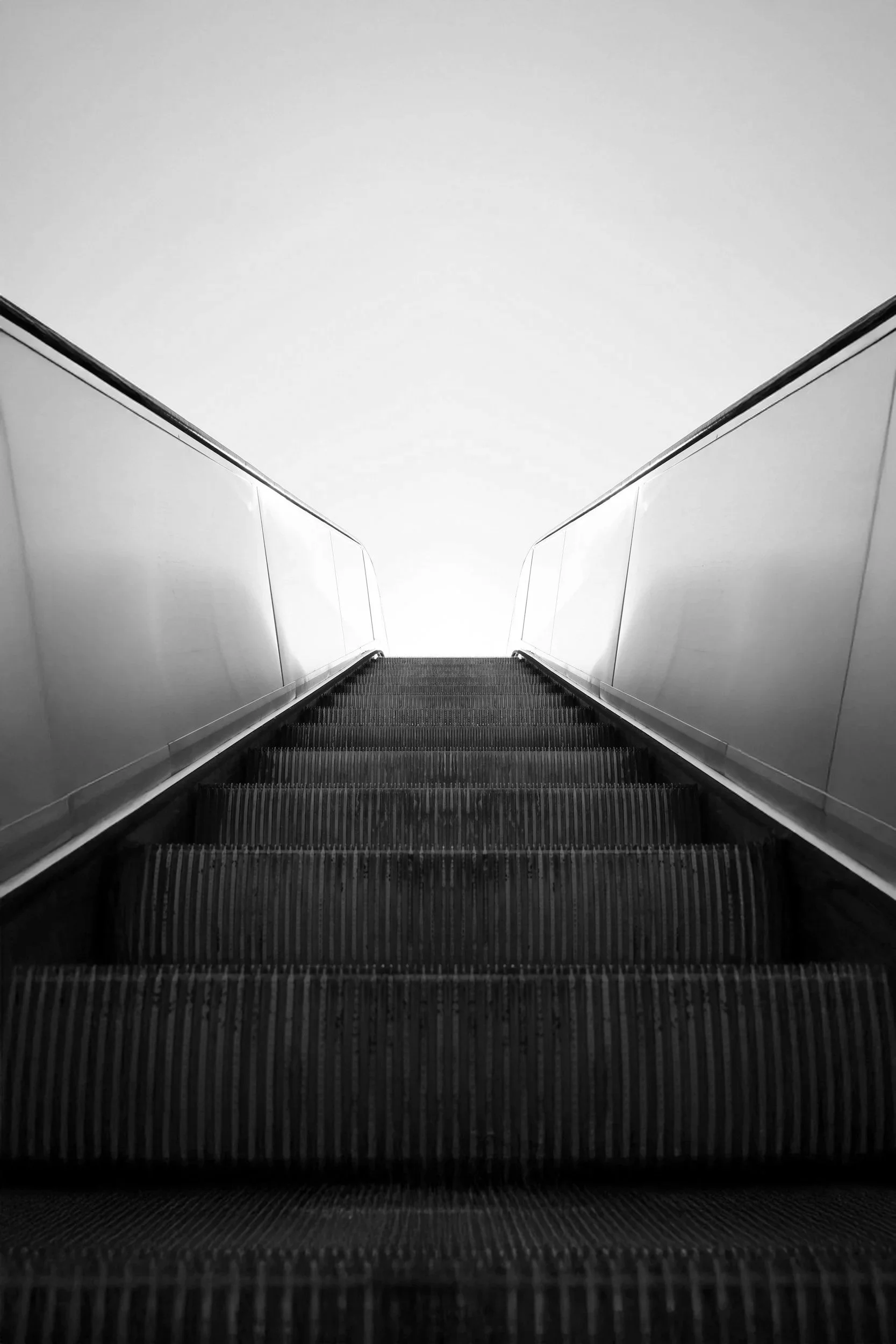 Black and white photo of an upward view of an escalator with handrails on both sides, leading towards the bright sky.