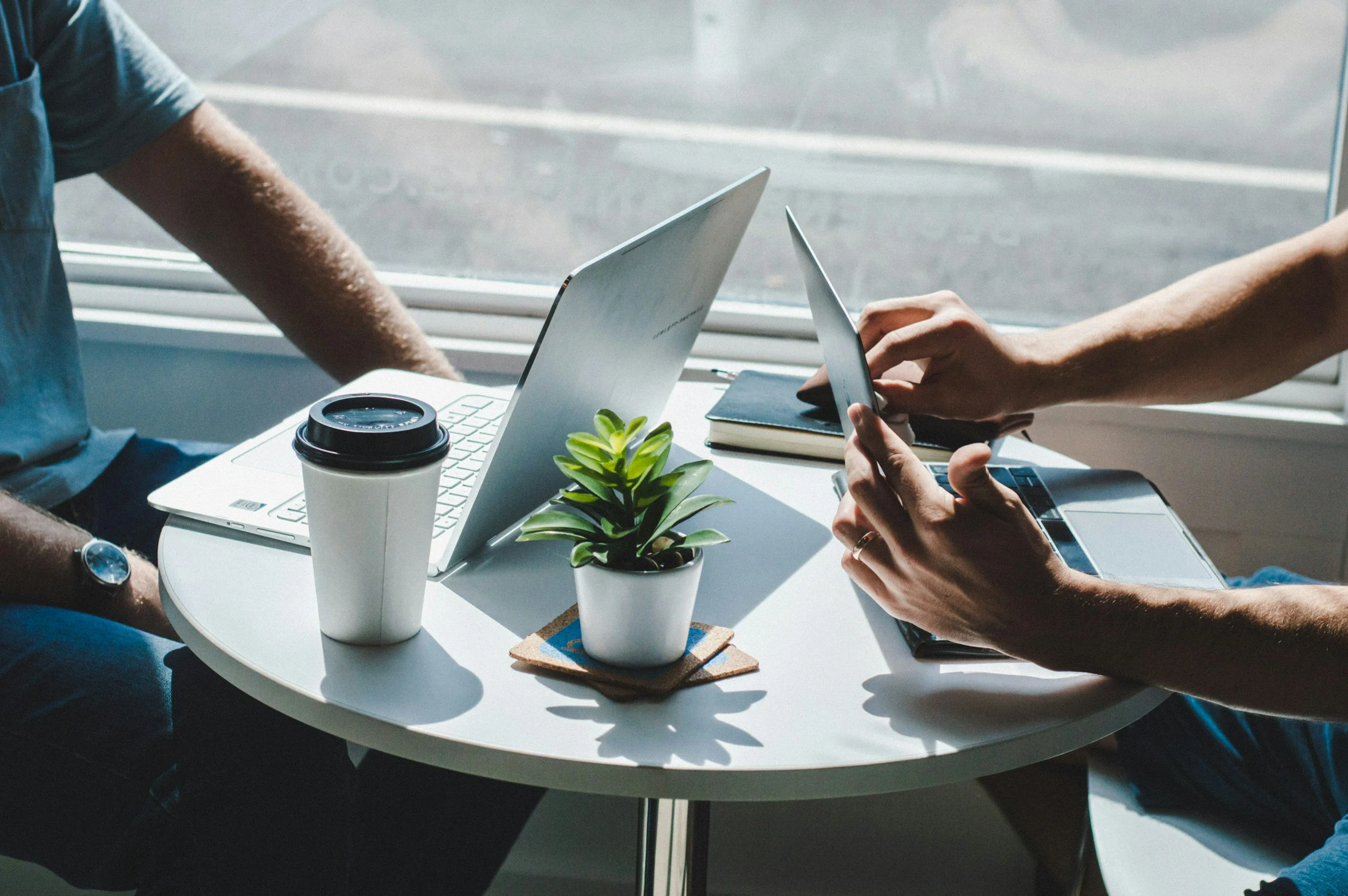 Two people sitting at a small round white table with laptops, a potted green plant, a black notebook, a paper coffee cup, and a smartphone.