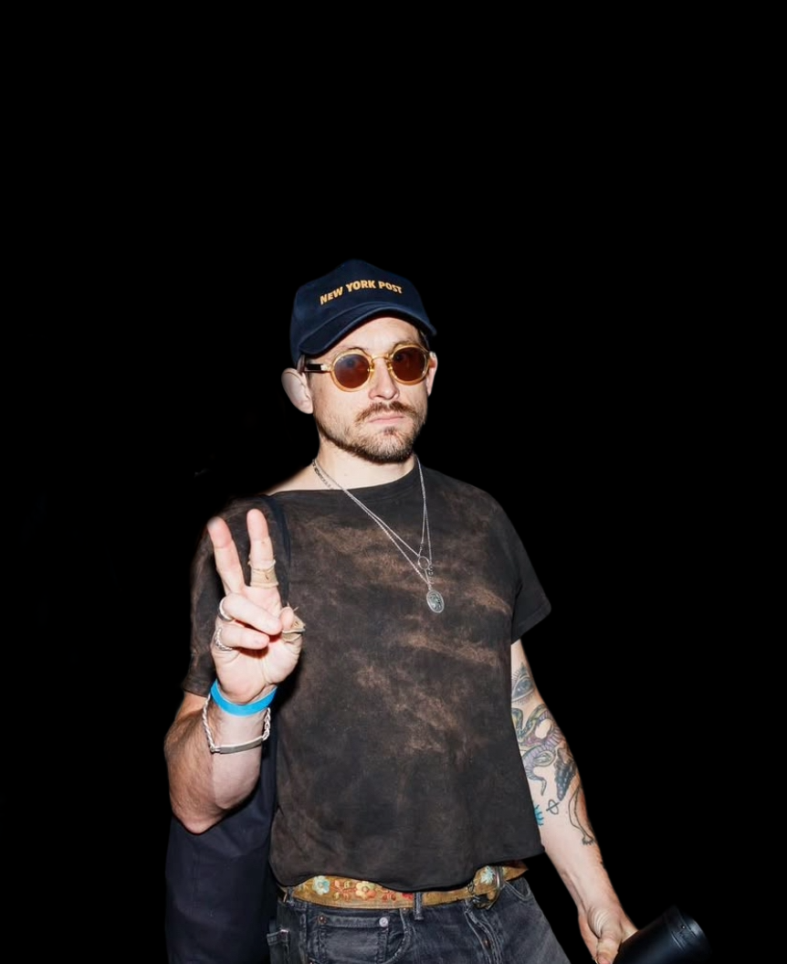A man with a beard and sunglasses making a peace sign, wearing a black cap with 'New York Post' written on it, a black torn shirt, and various jewelry, standing against a black background.