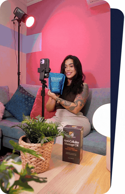 A woman with tattoos on her arms sitting on a gray couch, holding a blue box labeled 'Ancient Remedies,' with a smile. There is a beauty product box labeled 'Anti-Cellulite Massage Oil' on the table in front of her. She is being filmed with a smartphone on a tripod, and a red studio light is shining nearby, in a pink indoor setting.