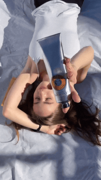 A woman lying on a bed, holding a tube of skincare or cosmetic product in front of her face.