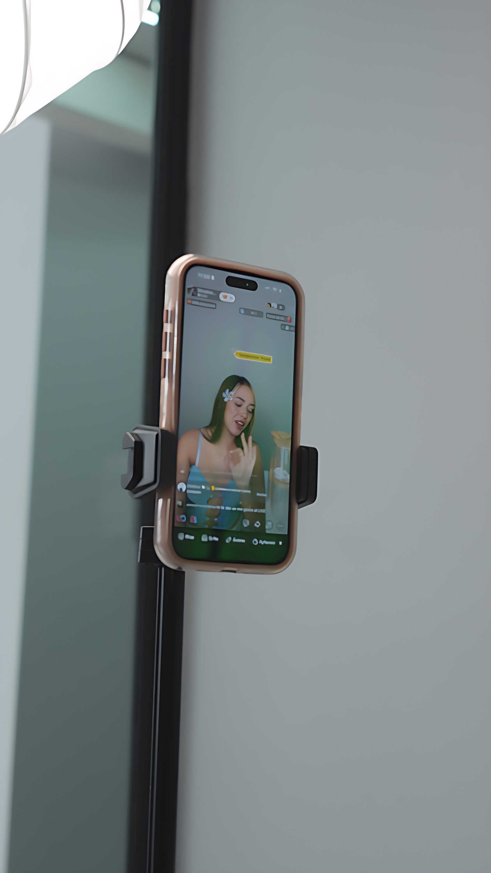 A smartphone mounted on a refrigerator door is recording a woman with green hair, a flower hair clip, and a blue top, who appears to be singing or speaking.