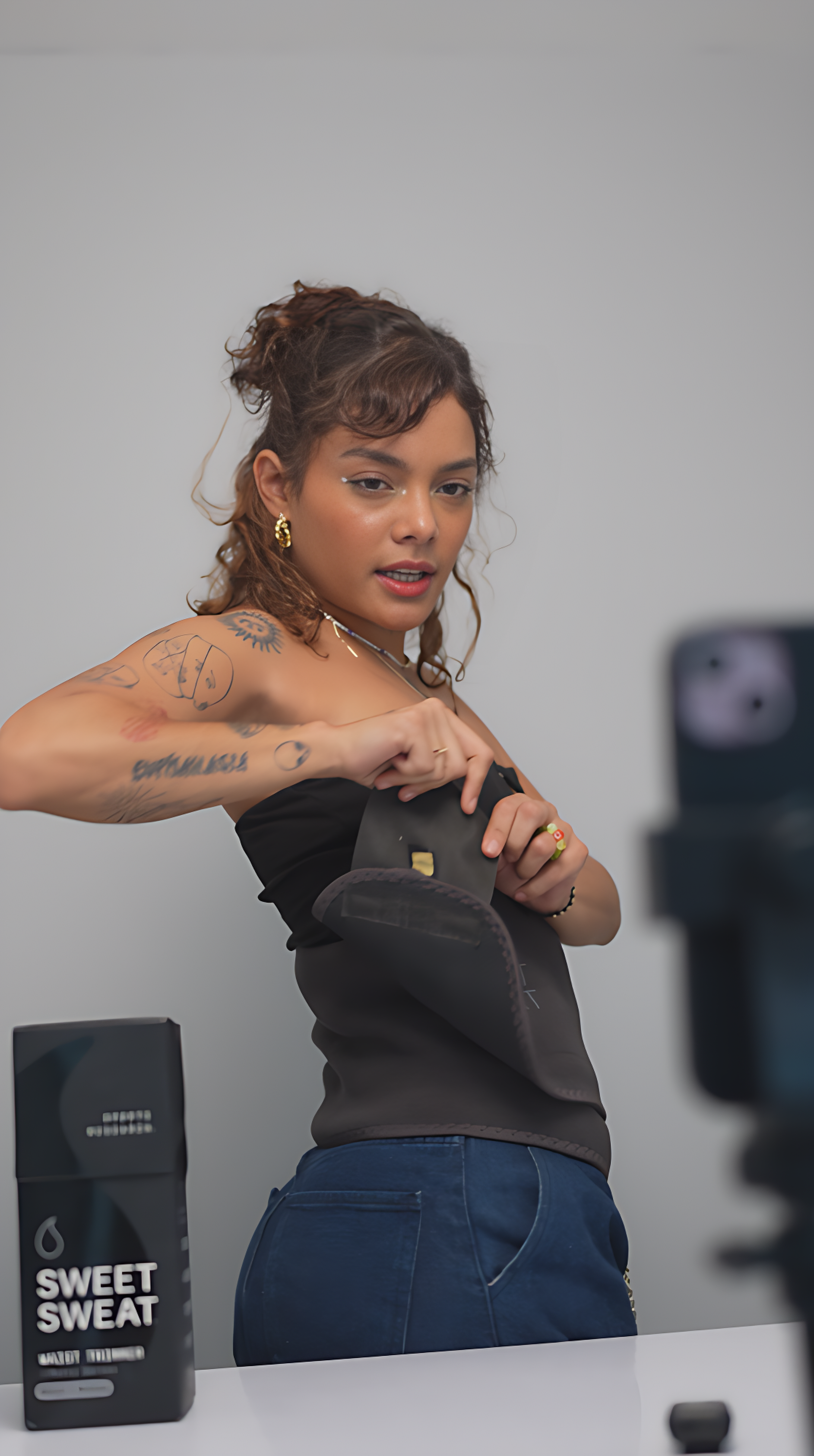 Young woman with tattoos and earrings, wearing a black top, preparing or adjusting a black item, standing in a room with a white wall, with a box labeled 'Sweet Sweat' on a table in the background, and a camera capturing her image.