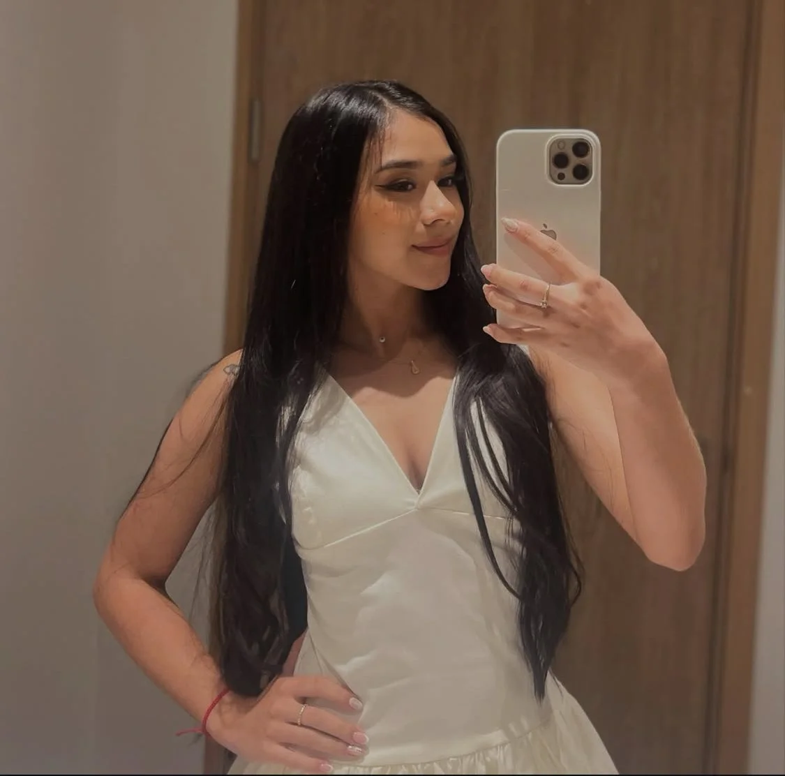 Young woman with long dark hair taking a mirror selfie with her iPhone, wearing a white dress, in front of a wooden door.