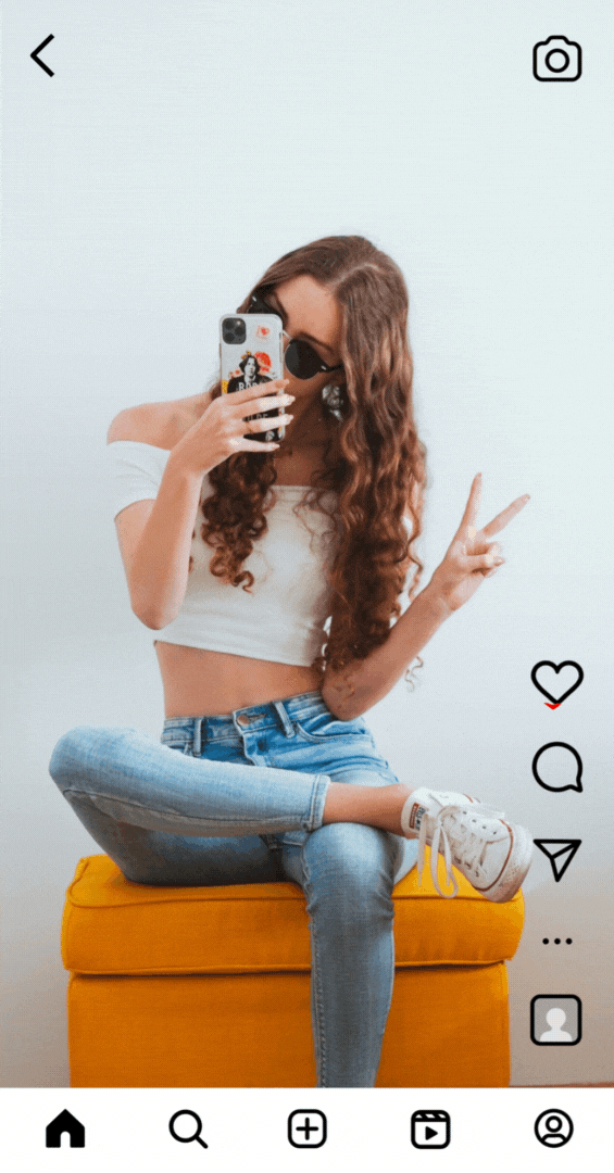 Young woman with long curly hair sitting cross-legged on a yellow ottoman, taking a selfie with her phone, wearing sunglasses, a white crop top, and jeans, making a peace sign with her right hand.
