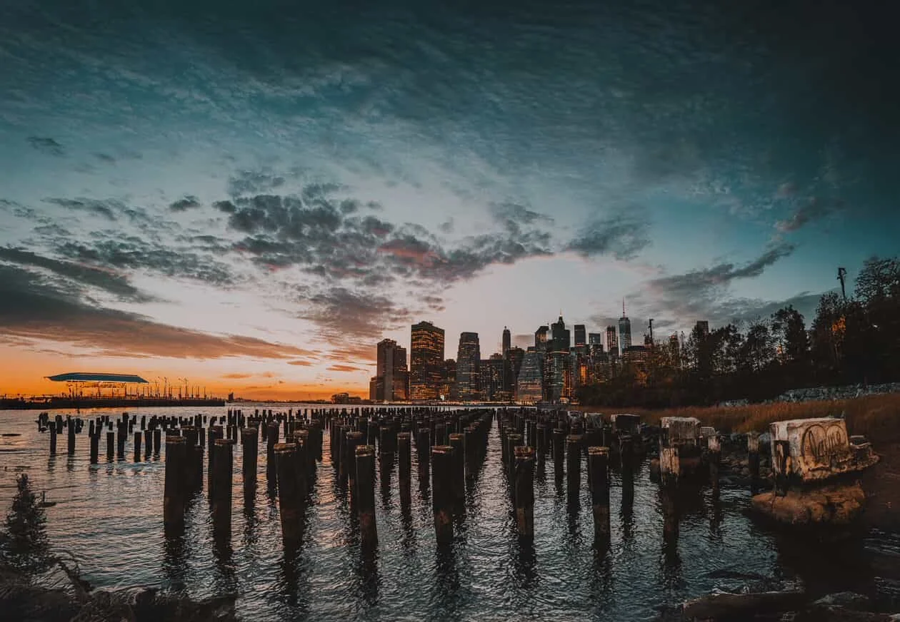 Sunset over a city skyline with water and old wooden pilings in the foreground.