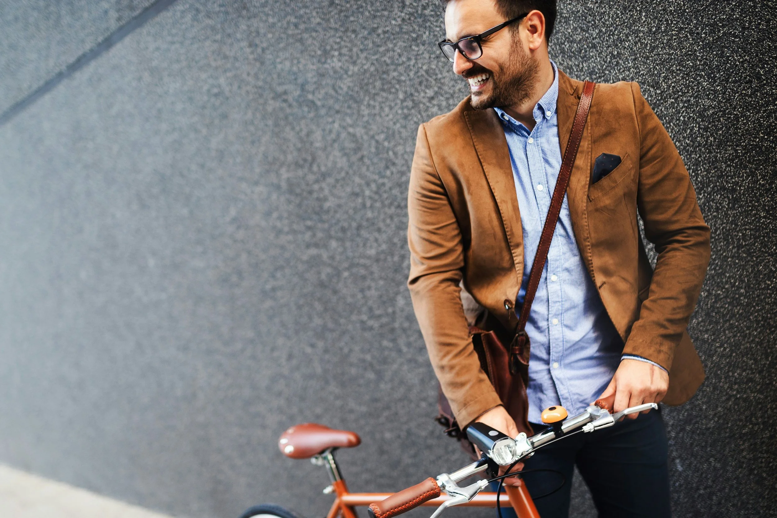 A man wearing glasses, a brown blazer, and a blue dress shirt smiling while holding the handlebars of a bicycle.